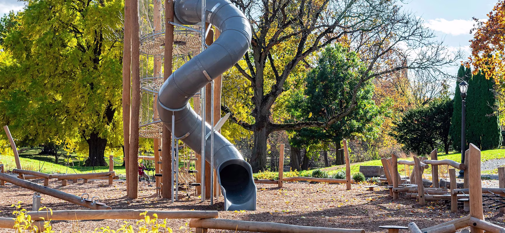 Empty playground with tall spiral tube slide and wooden climbing structures, set among large trees with bright autumn foliage