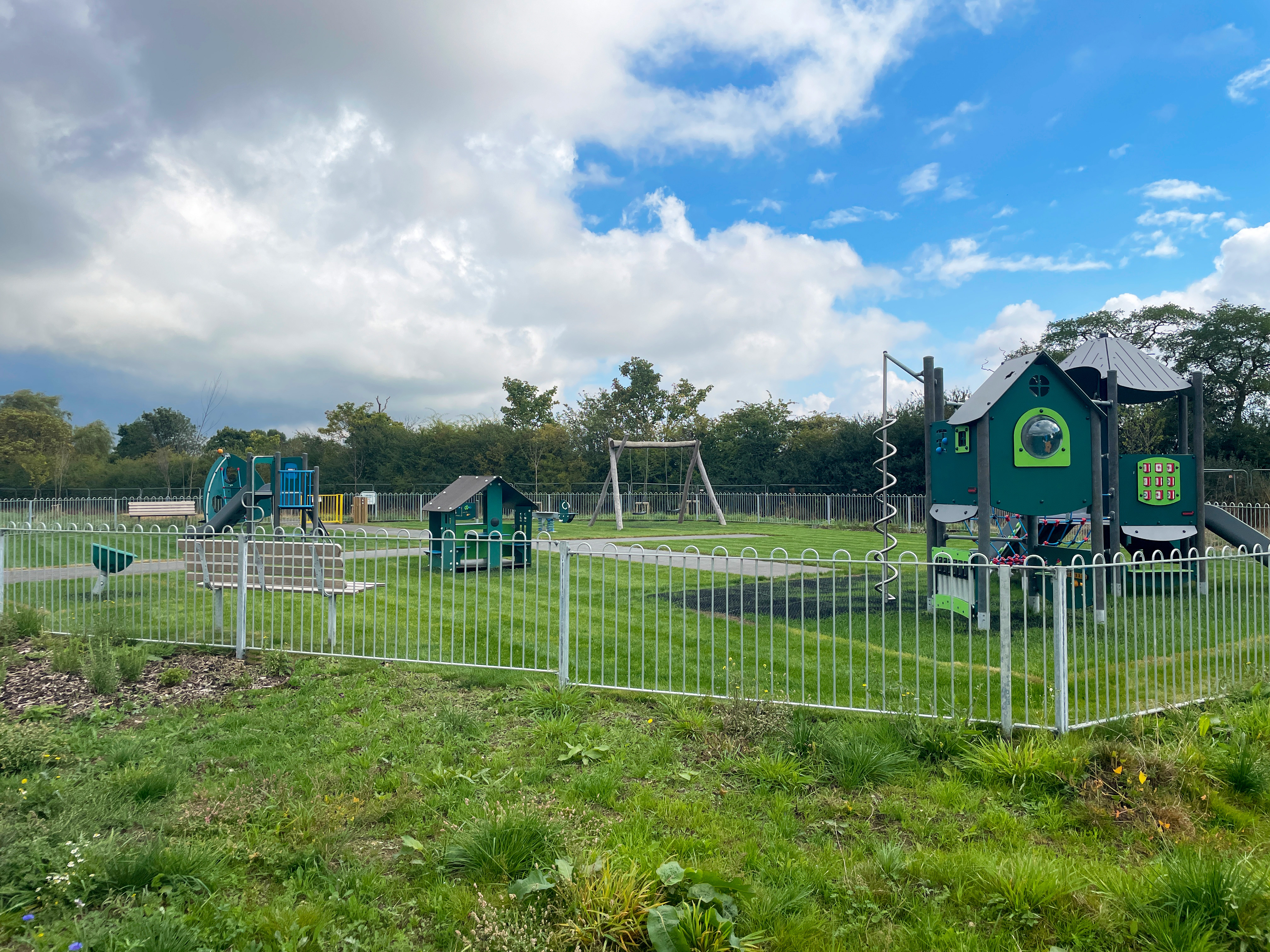 Fenced outdoor playground with green and blue play structures, swings and benches on bright grass under a cloudy blue sky