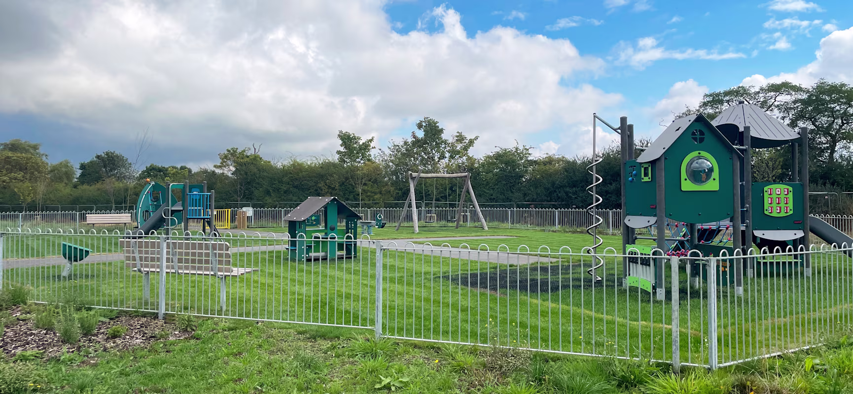 Fenced outdoor playground with green and blue play structures, swings and benches on bright grass under a cloudy blue sky