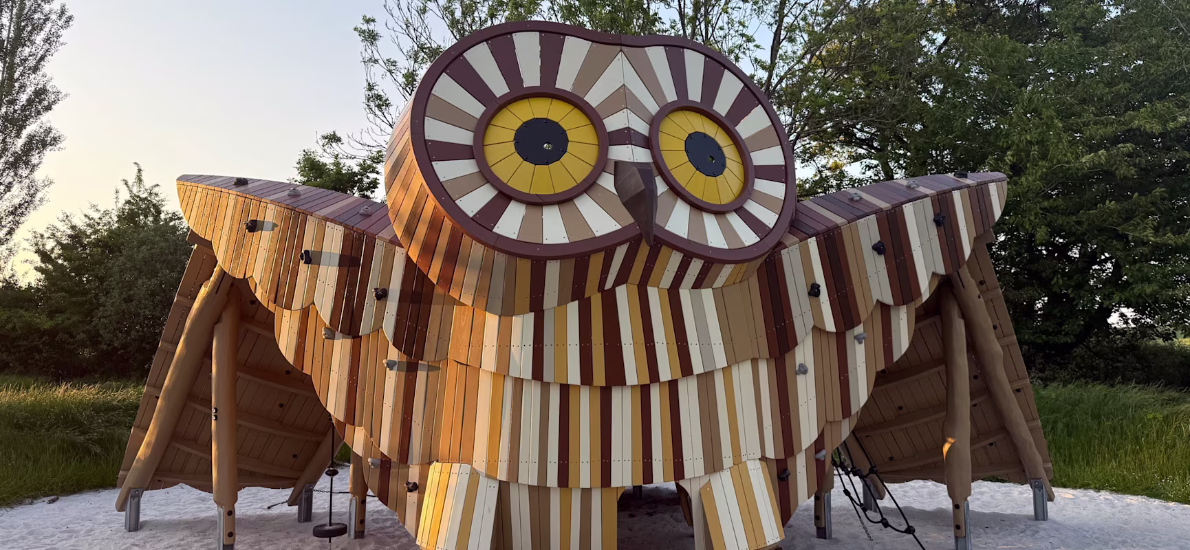Large striped wooden owl-shaped playground structure on sand, set in a grassy park with trees under a clear evening sky