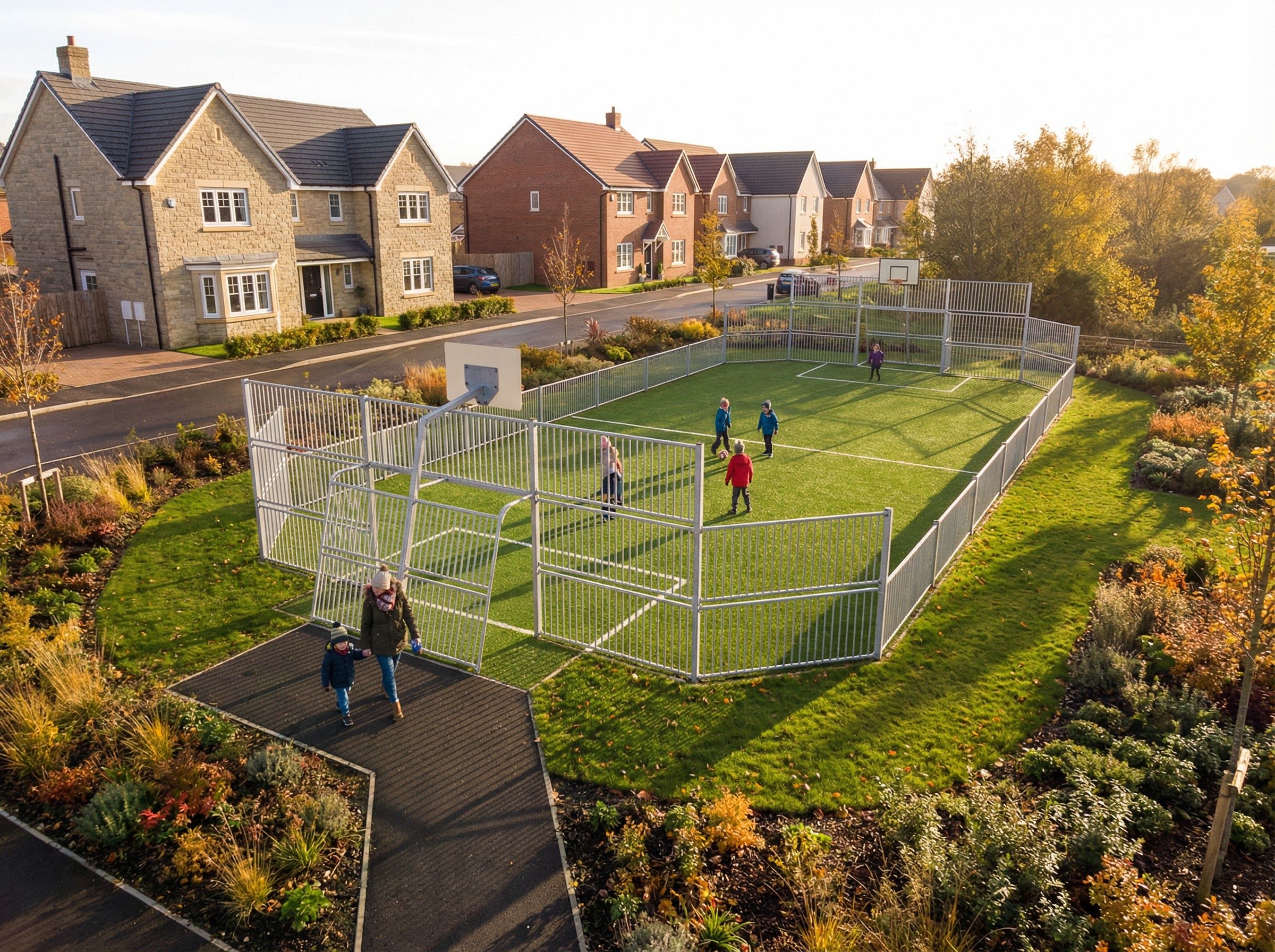 Children play on a fenced sports pitch beside a landscaped path and modern houses on a sunny day in a suburban neighbourhood
