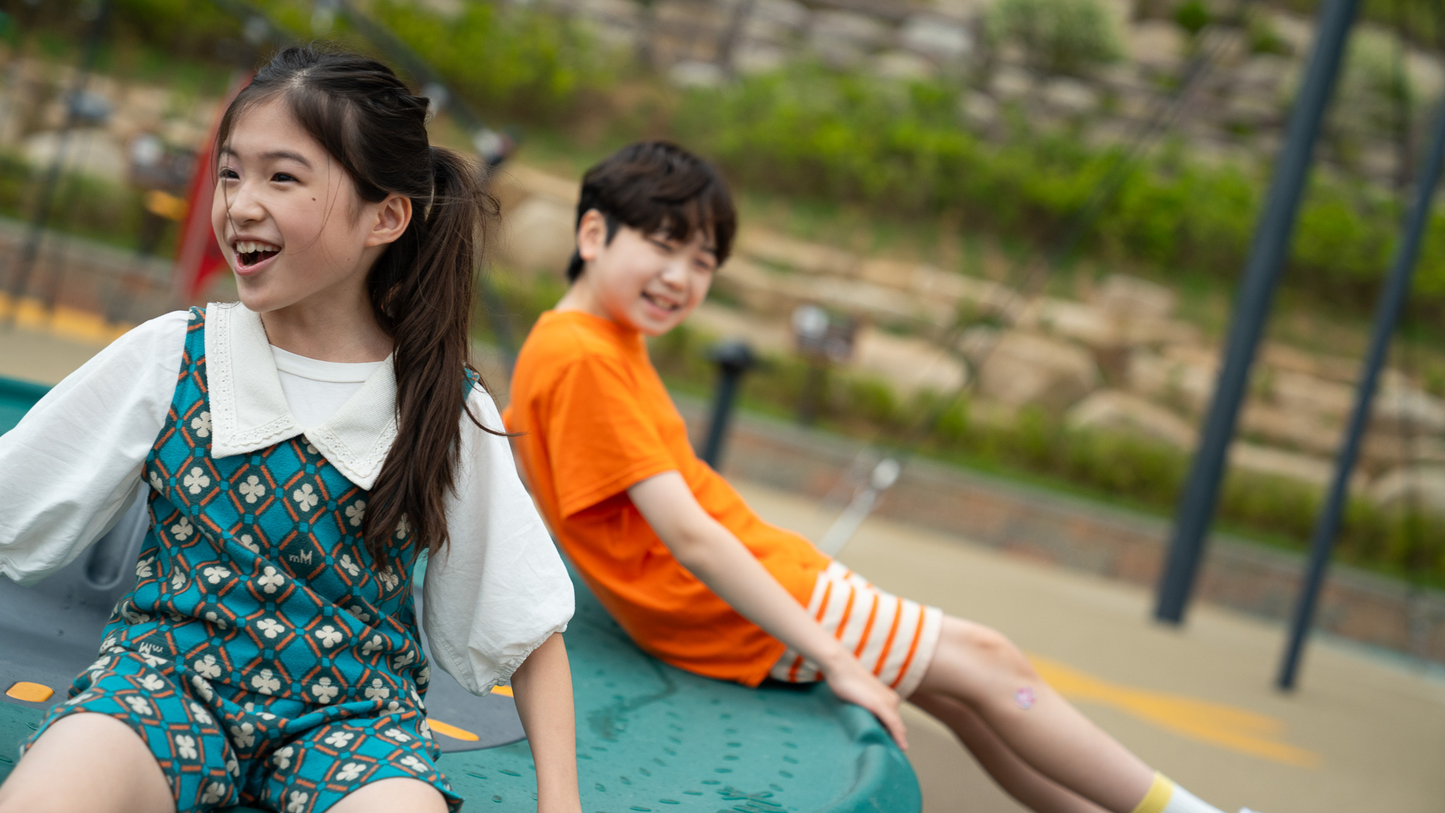Two children with blurred faces recline on playground equipment outdoors, one in a patterned teal outfit, the other in orange.