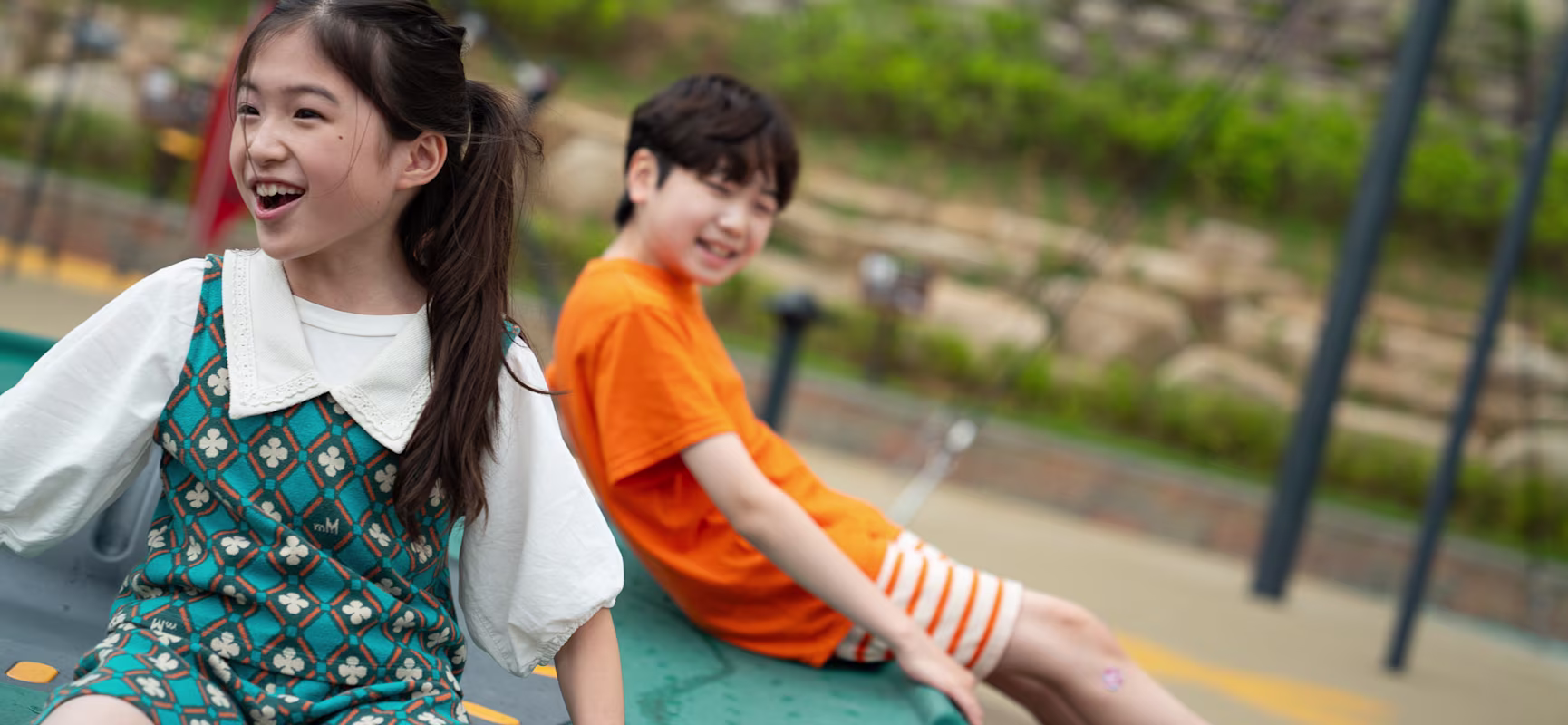 Two children with blurred faces recline on playground equipment outdoors, one in a patterned teal outfit, the other in orange.