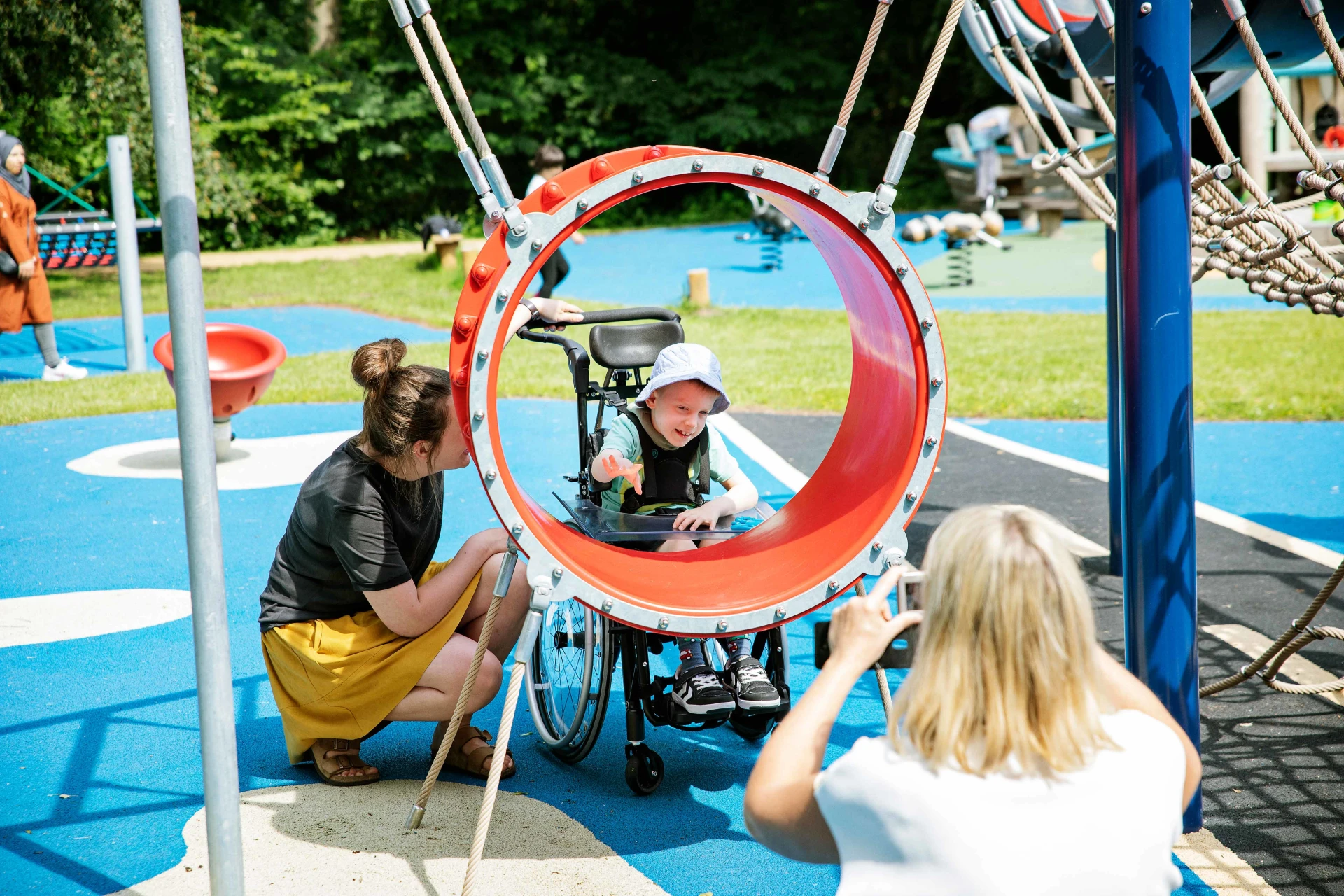 eine Frau fotografiert einen Jungen im Rollstuhl, der auf einem Spielplatz spielt