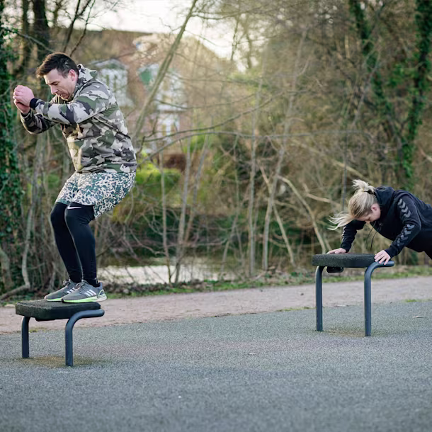Two people exercise outdoors, one jumping onto a platform, the other doing incline push-ups on another platform in a park setting.