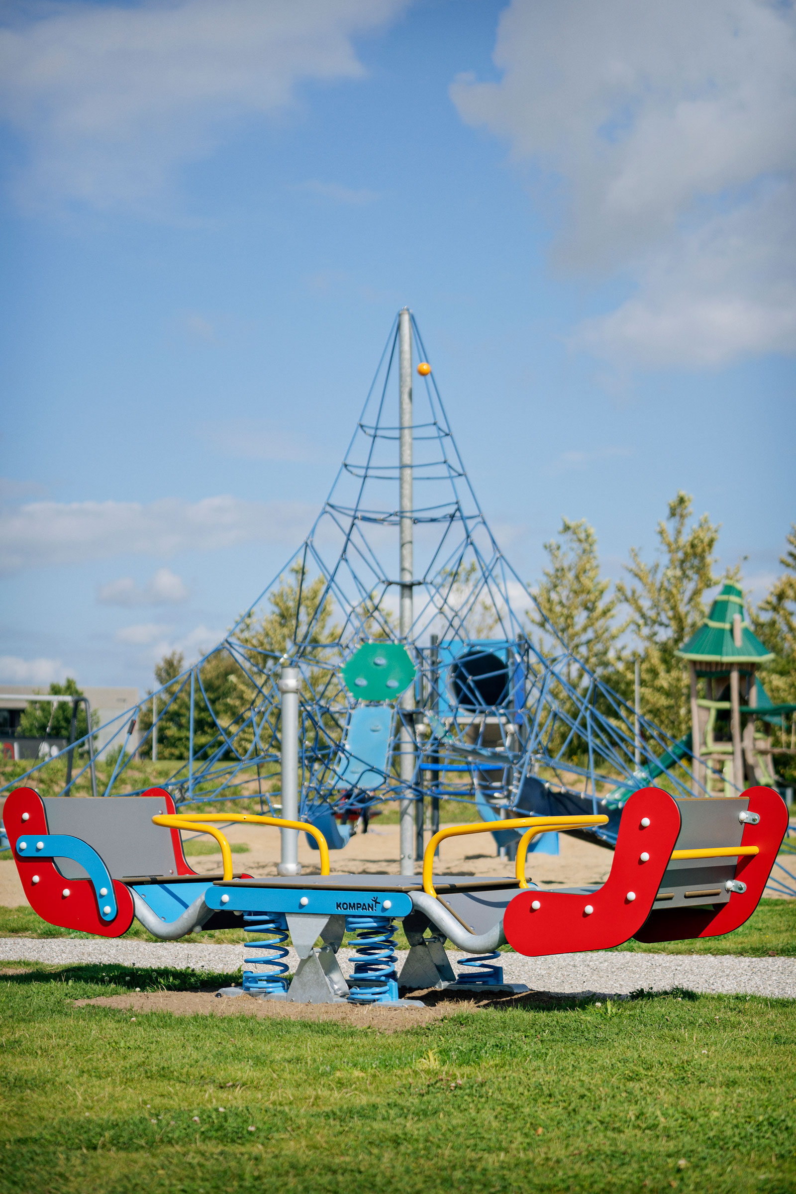 Colorful spring rocker and climbing net structure in a sunny outdoor playground with grass, sand, and trees in the background.