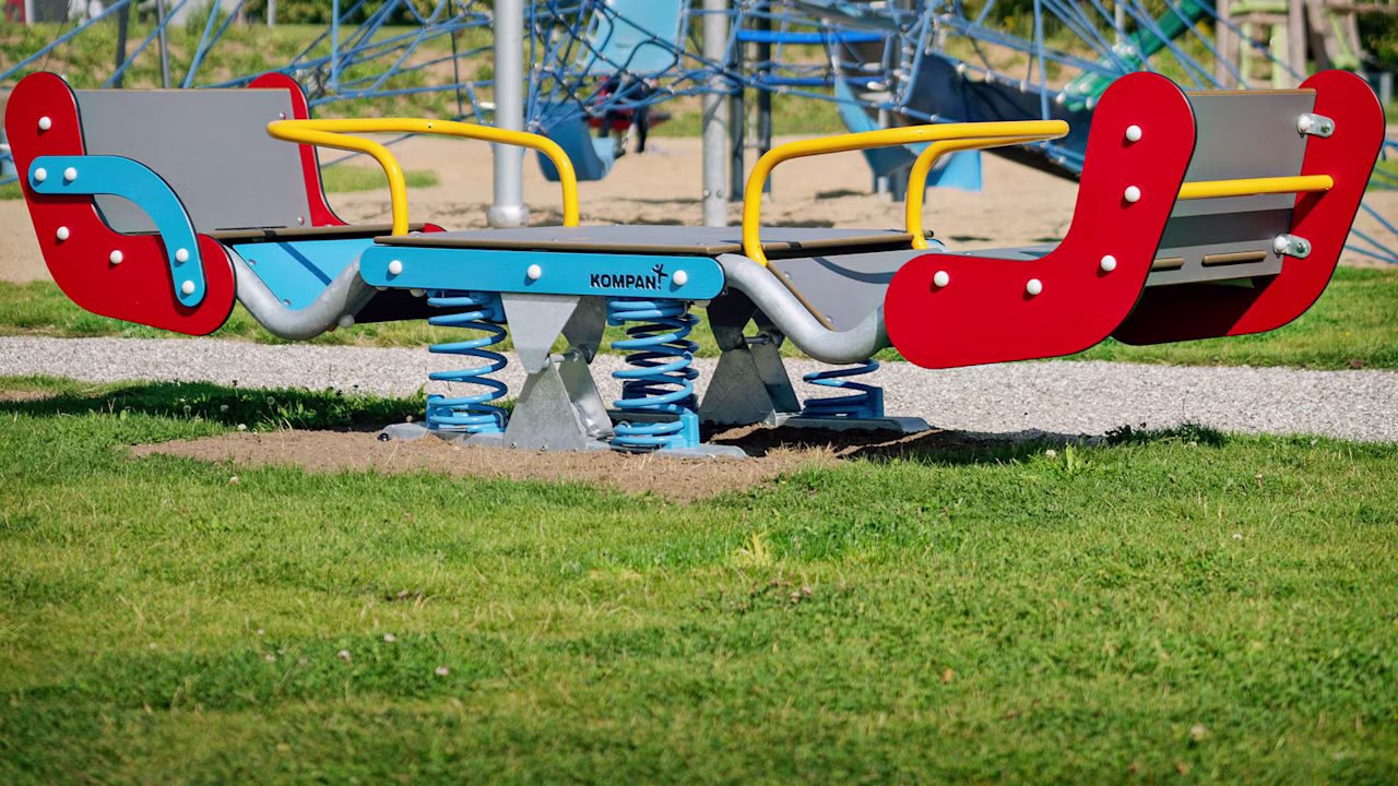 Colorful spring rocker and climbing net structure in a sunny outdoor playground with grass, sand, and trees in the background.