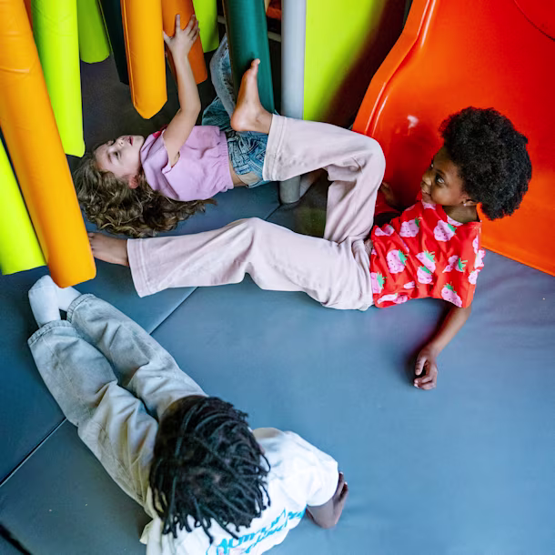 Three children play on padded floor by colorful soft tubes and an orange slide, lying and climbing in an indoor play area