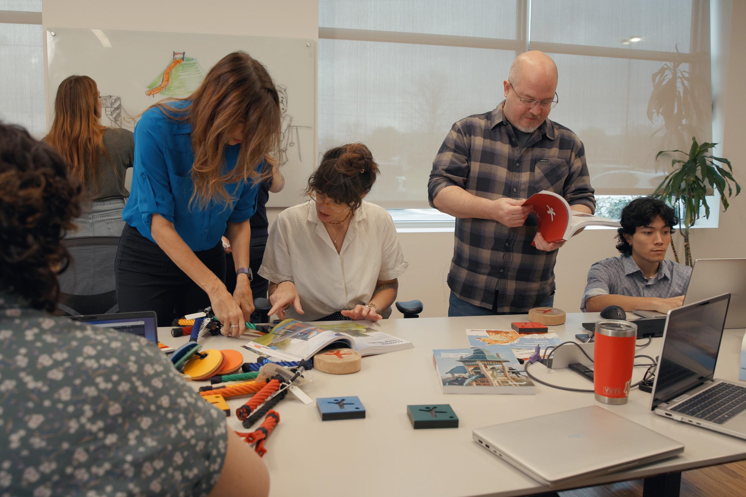 Team collaborating around a table with prototypes, books, and laptops, while one person sketches designs on a whiteboard in the background