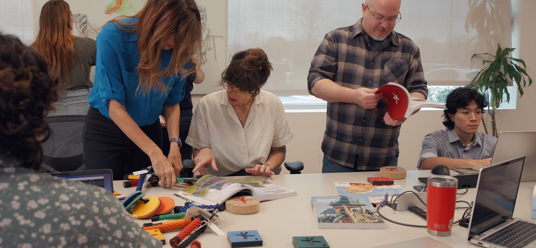 Team collaborating around a table with prototypes, books, and laptops, while one person sketches designs on a whiteboard in the background