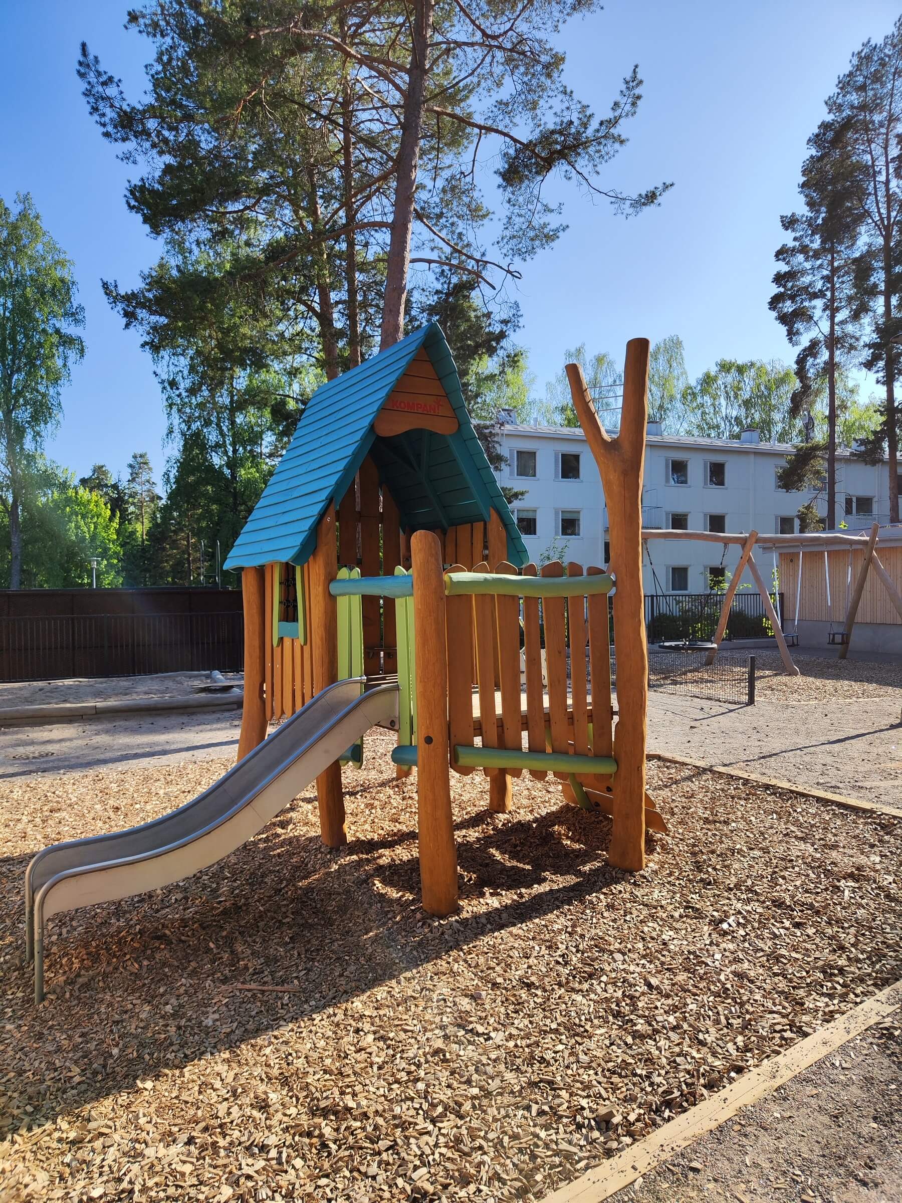 Wooden playground structure with blue roof and slide on wood chips, surrounded by trees and a residential building in the background