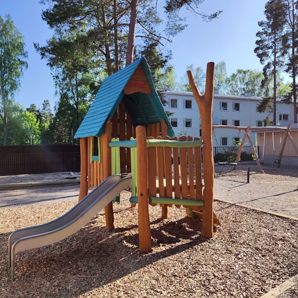 Wooden playground structure with blue roof and slide on wood chips, surrounded by trees and a residential building in the background