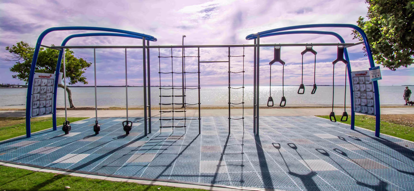 Outdoor fitness playground with blue frames, ropes and rings by a seaside path, under a cloudy sky with sunlight on the water