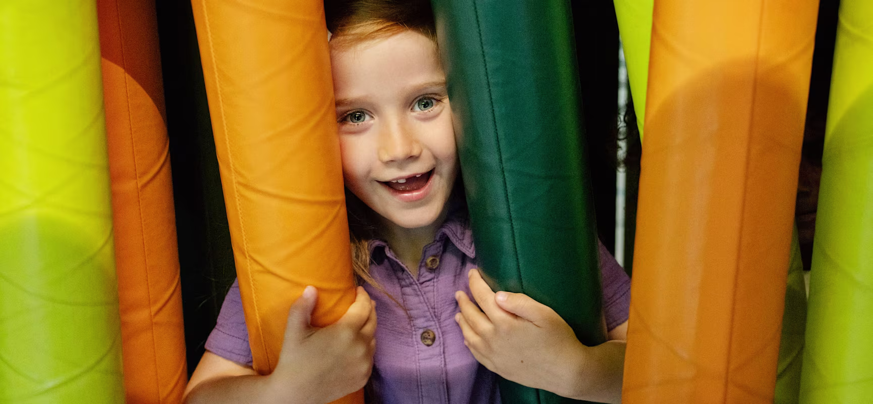Child in purple shirt standing among tall padded tubes in bright green, orange, and teal, holding them with both hands