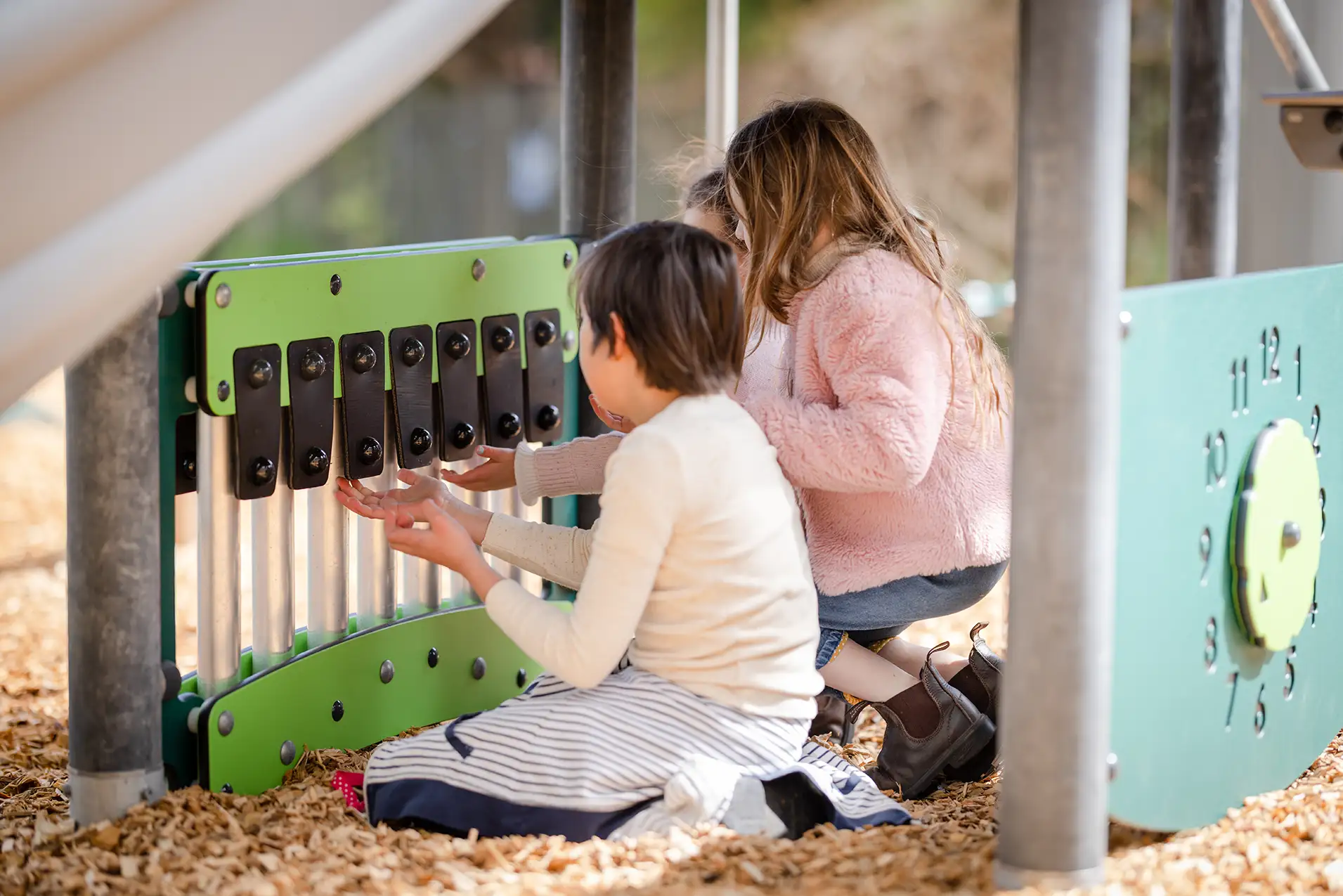 Children crouch at a playground music panel, playing silver chimes with black keys on green boards over woodchip ground