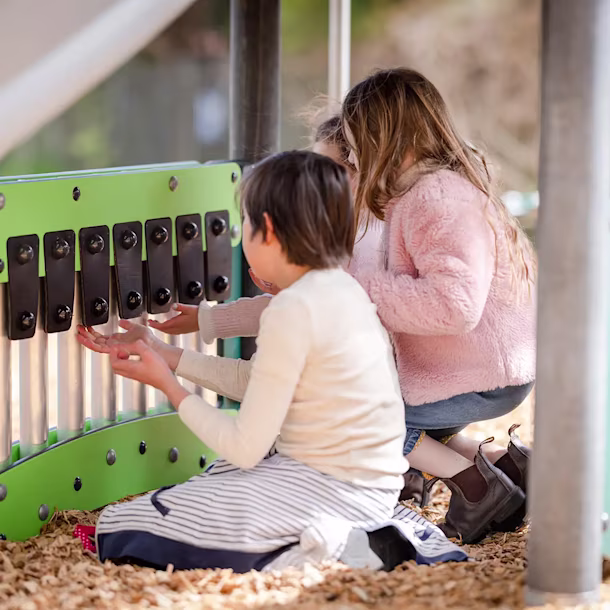 Children crouch at a playground music panel, playing silver chimes with black keys on green boards over woodchip ground