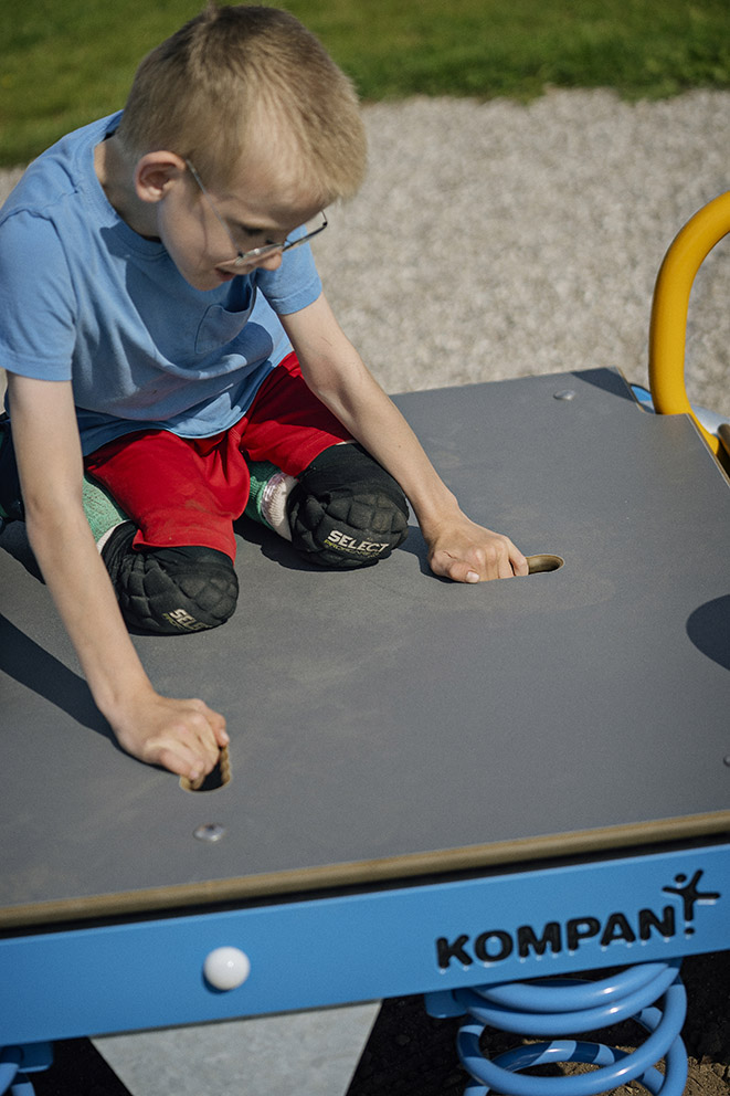 Child with blurred face kneels on a blue Kompan spring playground platform, gripping handholds while wearing red shorts and knee pads