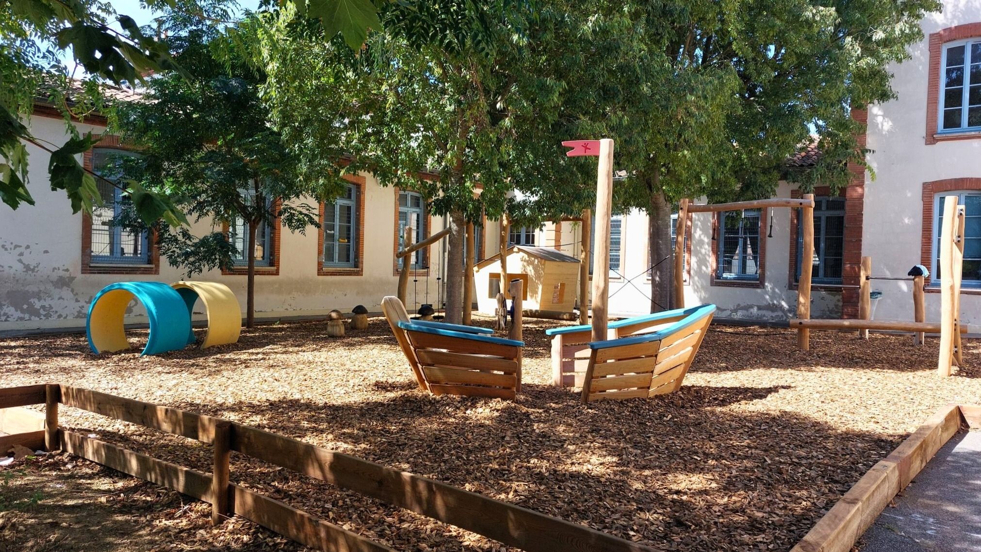 Sunny school playground with wood chips, boat-shaped climbing frames, colourful tunnels and trees in front of a building with blue windows