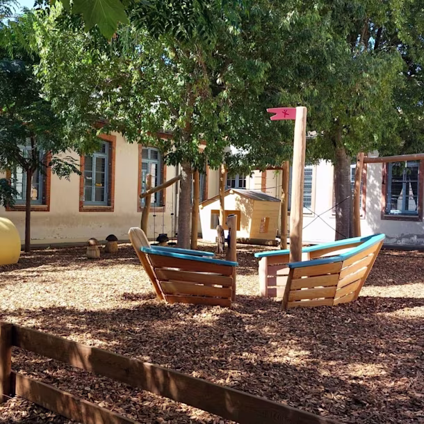 Sunny school playground with wood chips, boat-shaped climbing frames, colourful tunnels and trees in front of a building with blue windows