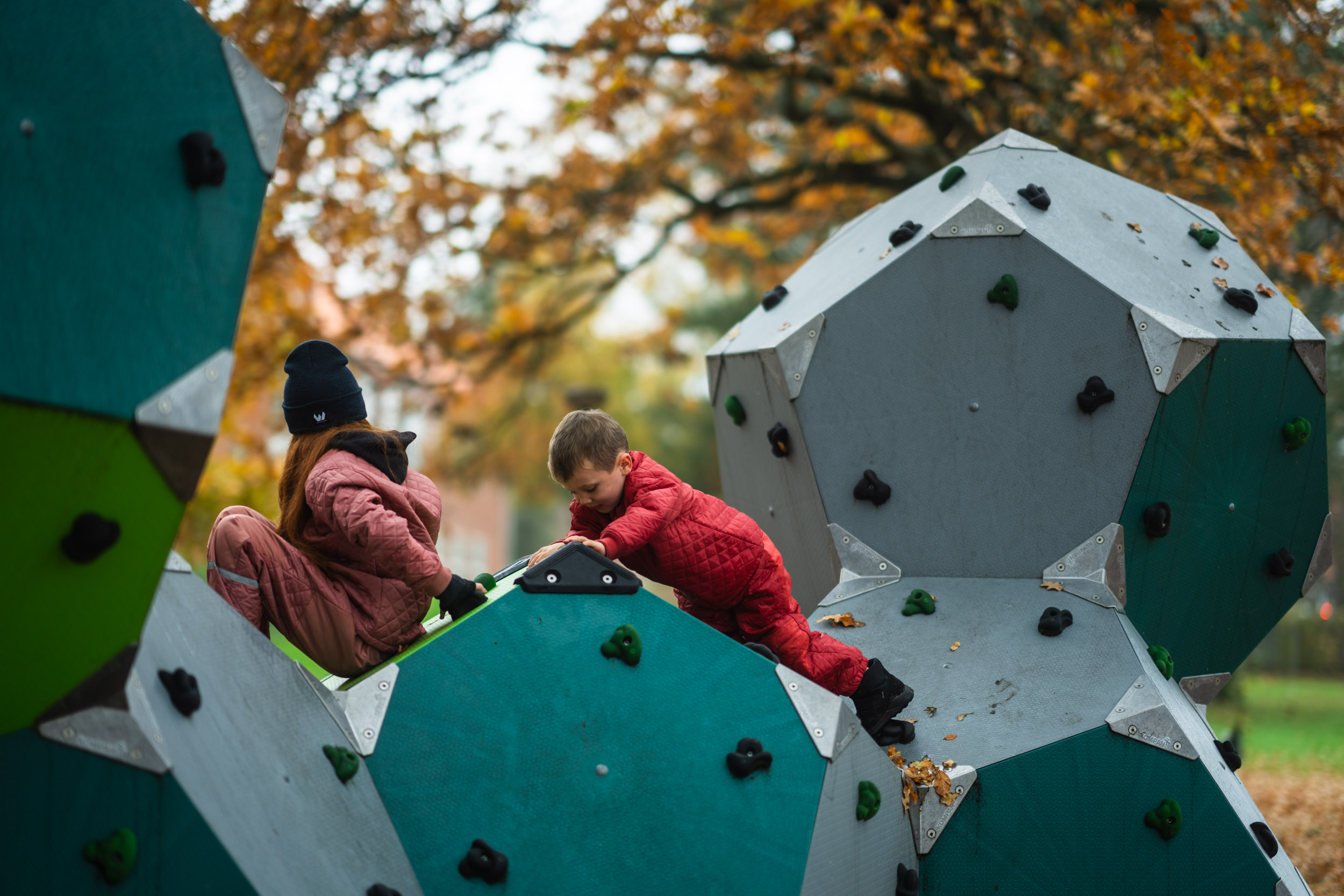Two children in red snowsuits climb a geometric climbing wall in a park, surrounded by autumn trees with orange leaves.