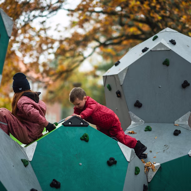 Two children in red snowsuits climb a geometric climbing wall in a park, surrounded by autumn trees with orange leaves.