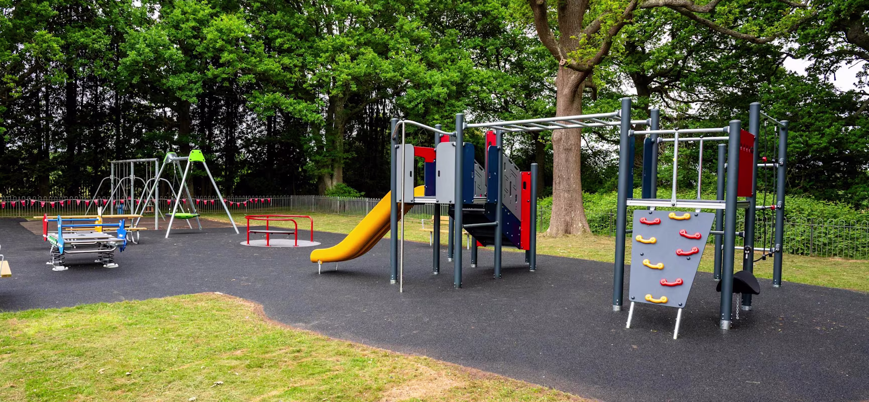 Empty outdoor playground with swings, climbing frames, and a yellow slide on black matting, surrounded by grass and tall green trees.