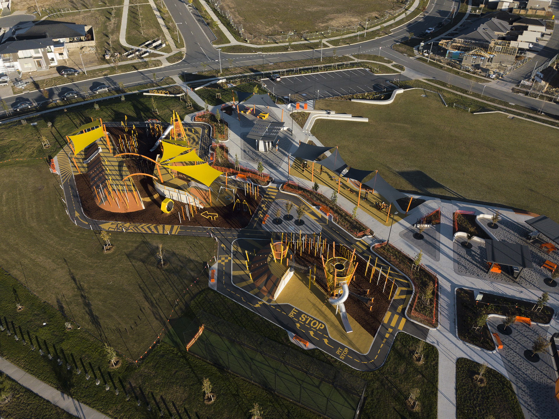 Aerial view of a modern playground with bright yellow structures, slides and shade sails, surrounded by roads, paths and open grass.