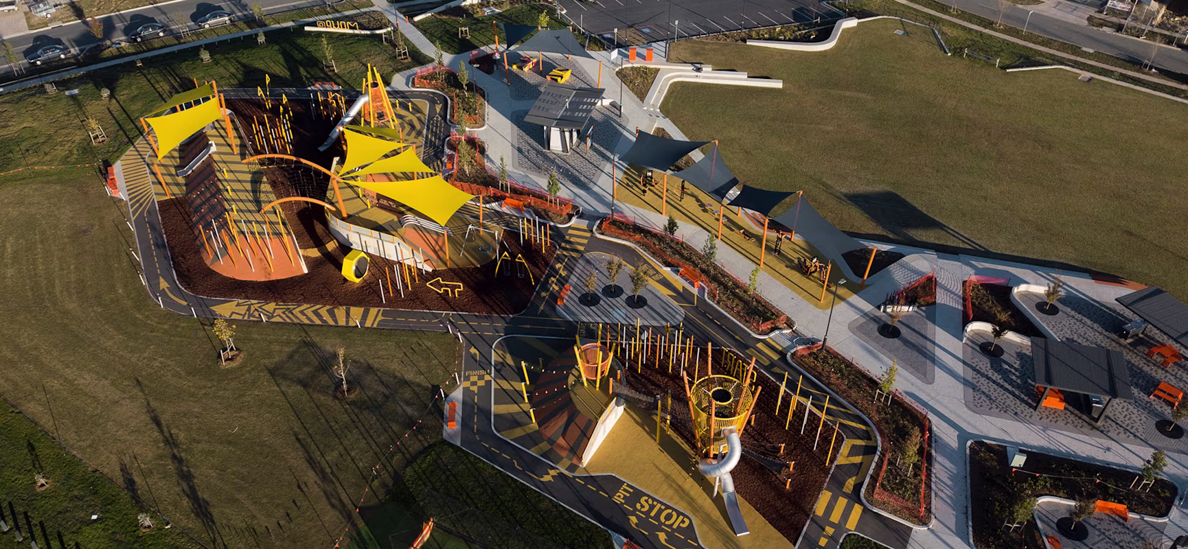 Aerial view of a modern playground with bright yellow structures, slides and shade sails, surrounded by roads, paths and open grass.