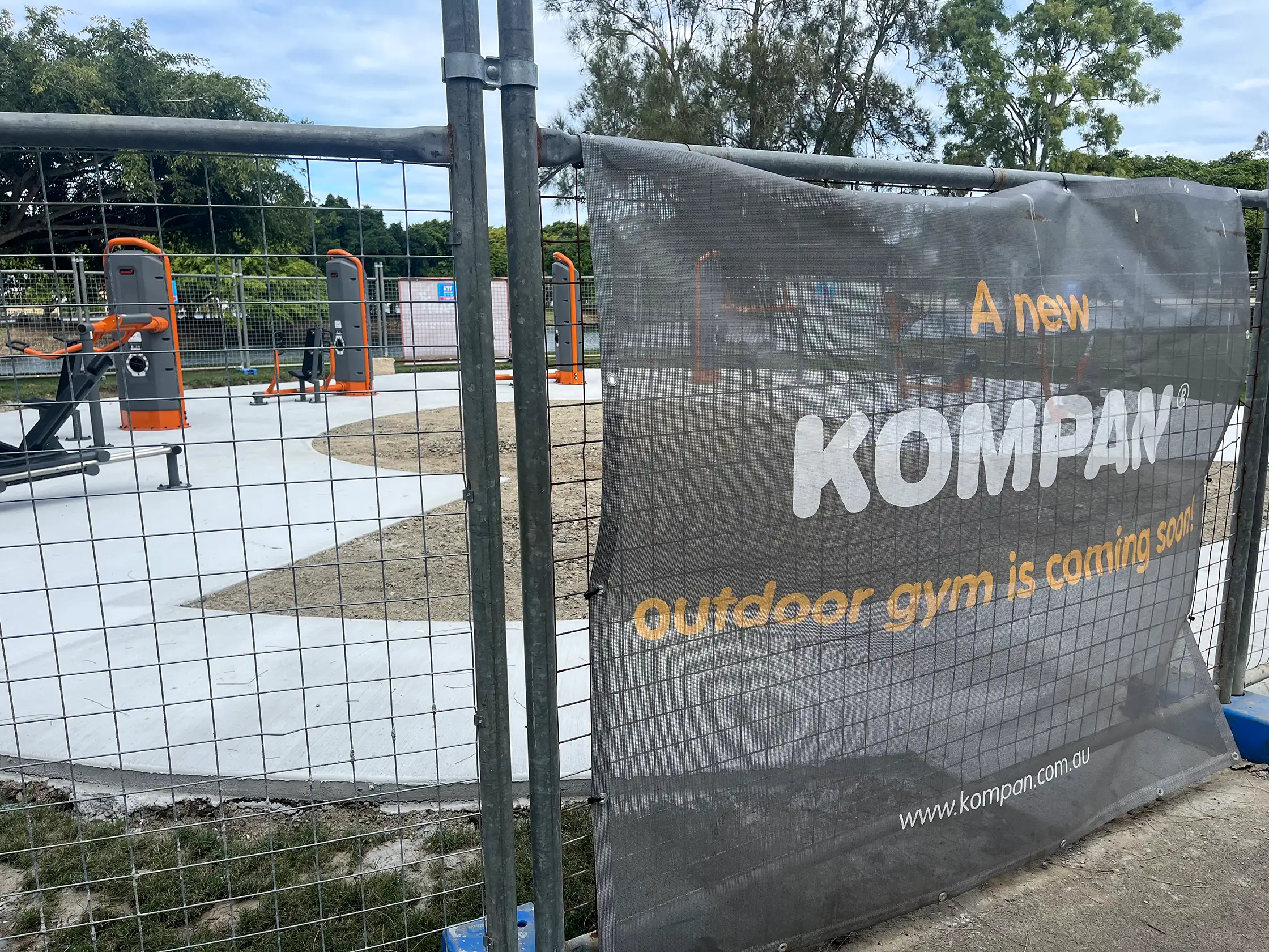 Fenced construction site with orange and grey outdoor gym equipment and a Kompan banner reading “A new outdoor gym is coming soon.”