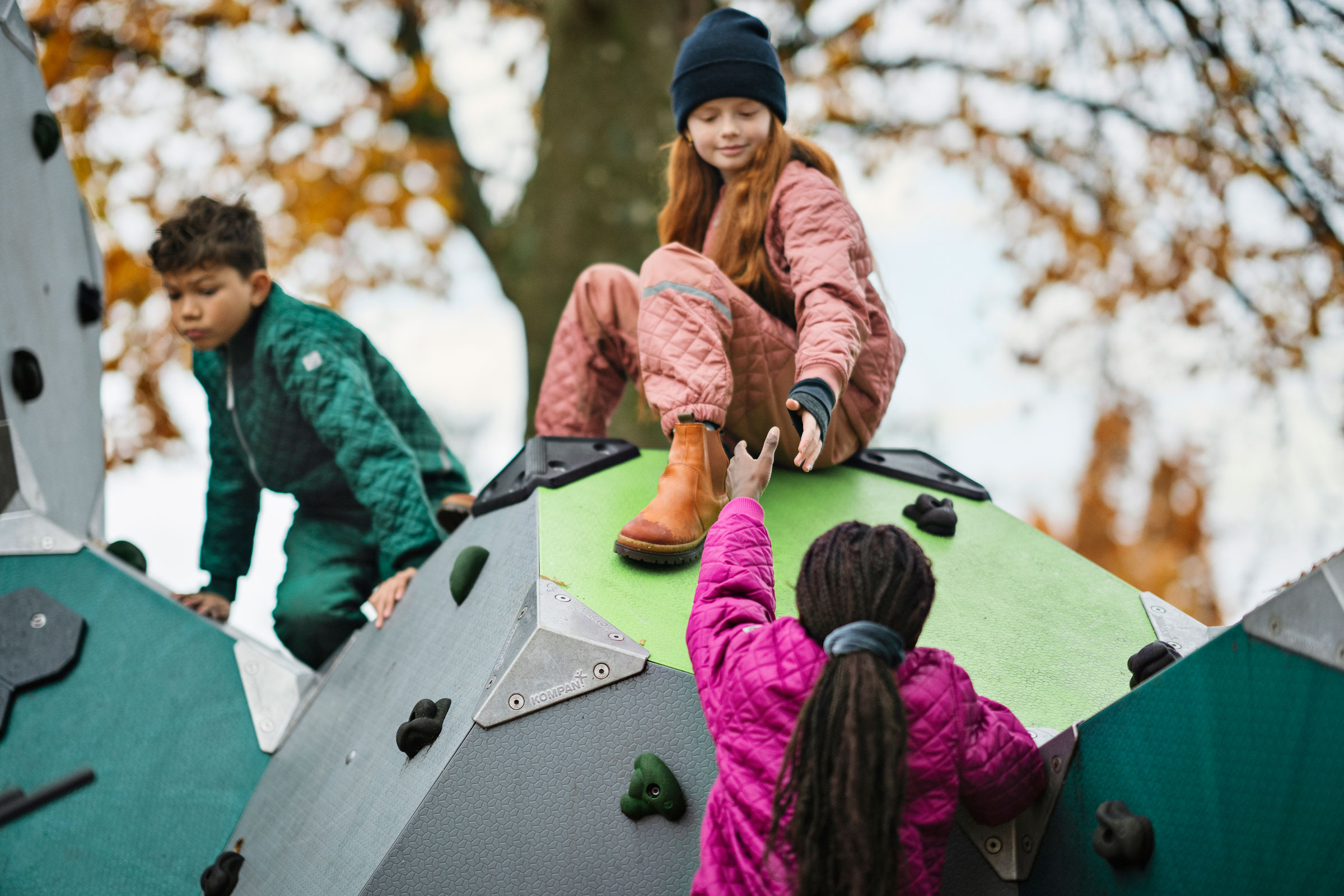 Children on a geometric climbing structure in autumn, one sitting on top reaching down to help another climb up