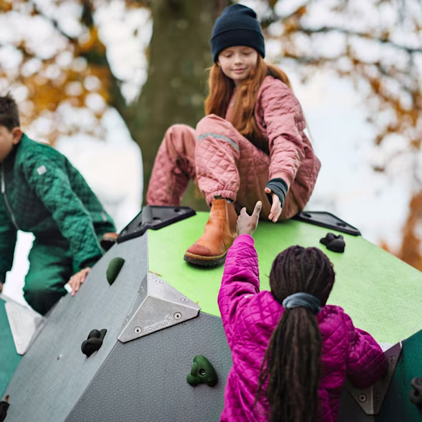 Children on a geometric climbing structure in autumn, one sitting on top reaching down to help another climb up