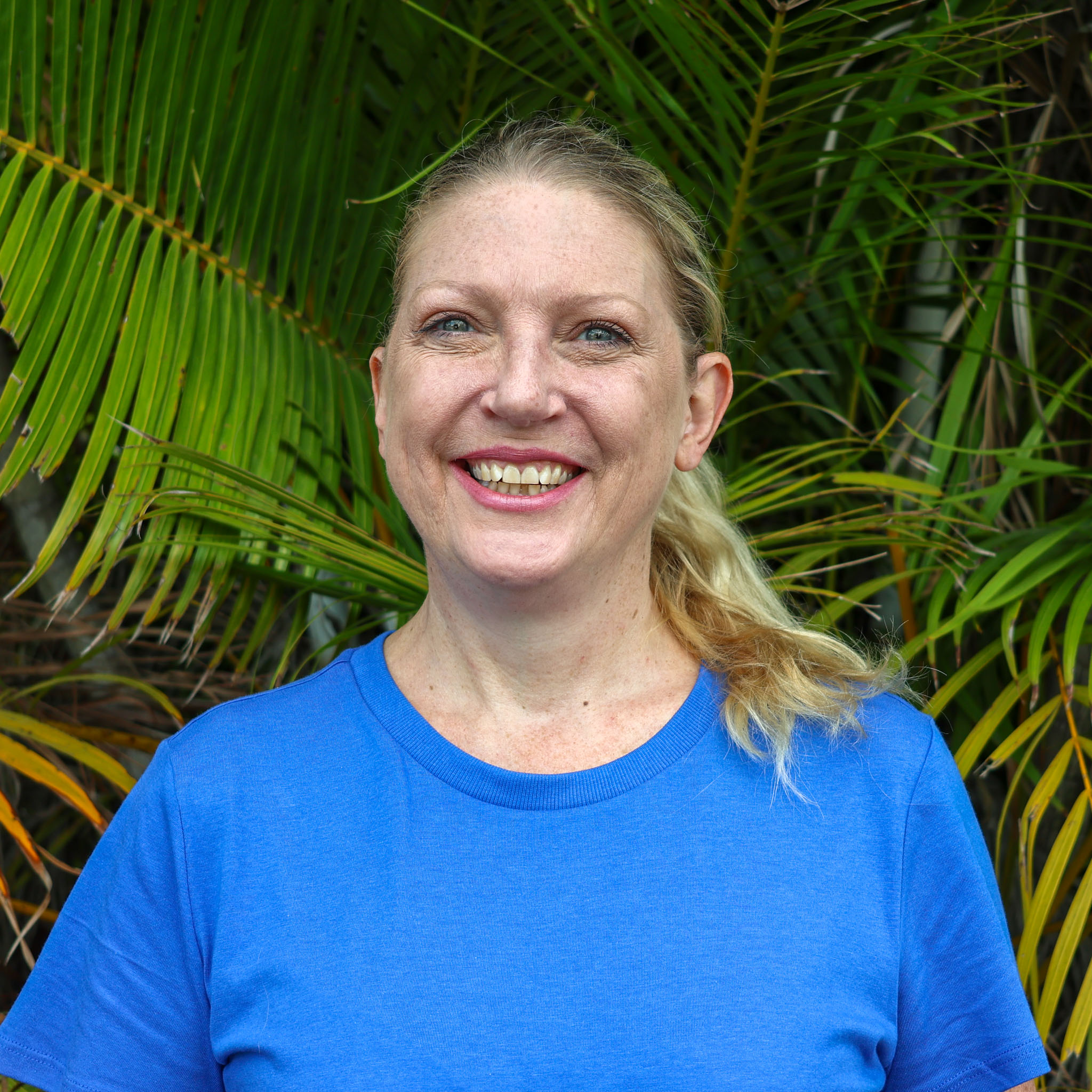 Person in a blue T-shirt standing in front of green palm leaves, with their face blurred for privacy