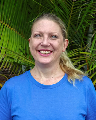 Person in a blue T-shirt standing in front of green palm leaves, with their face blurred for privacy