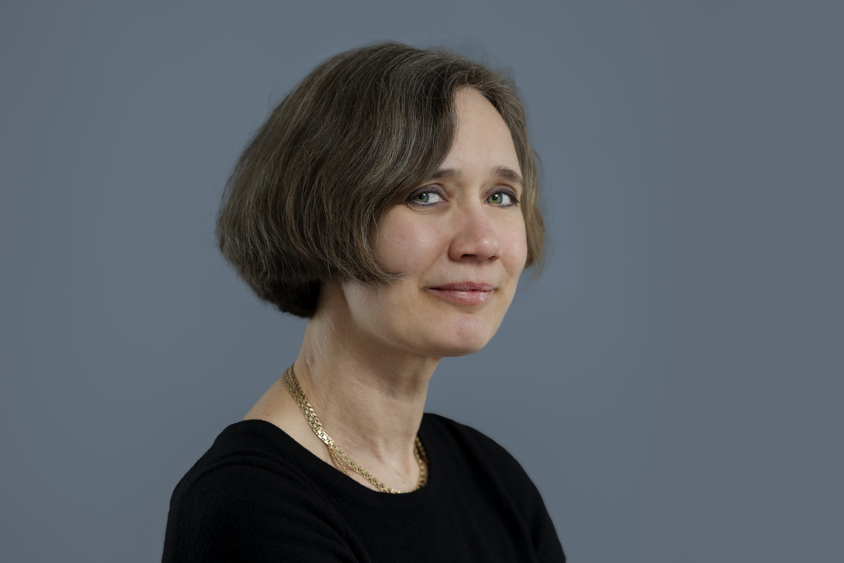Portrait of a person with blurred face, short brown hair, black top, and gold necklace against a plain grey background