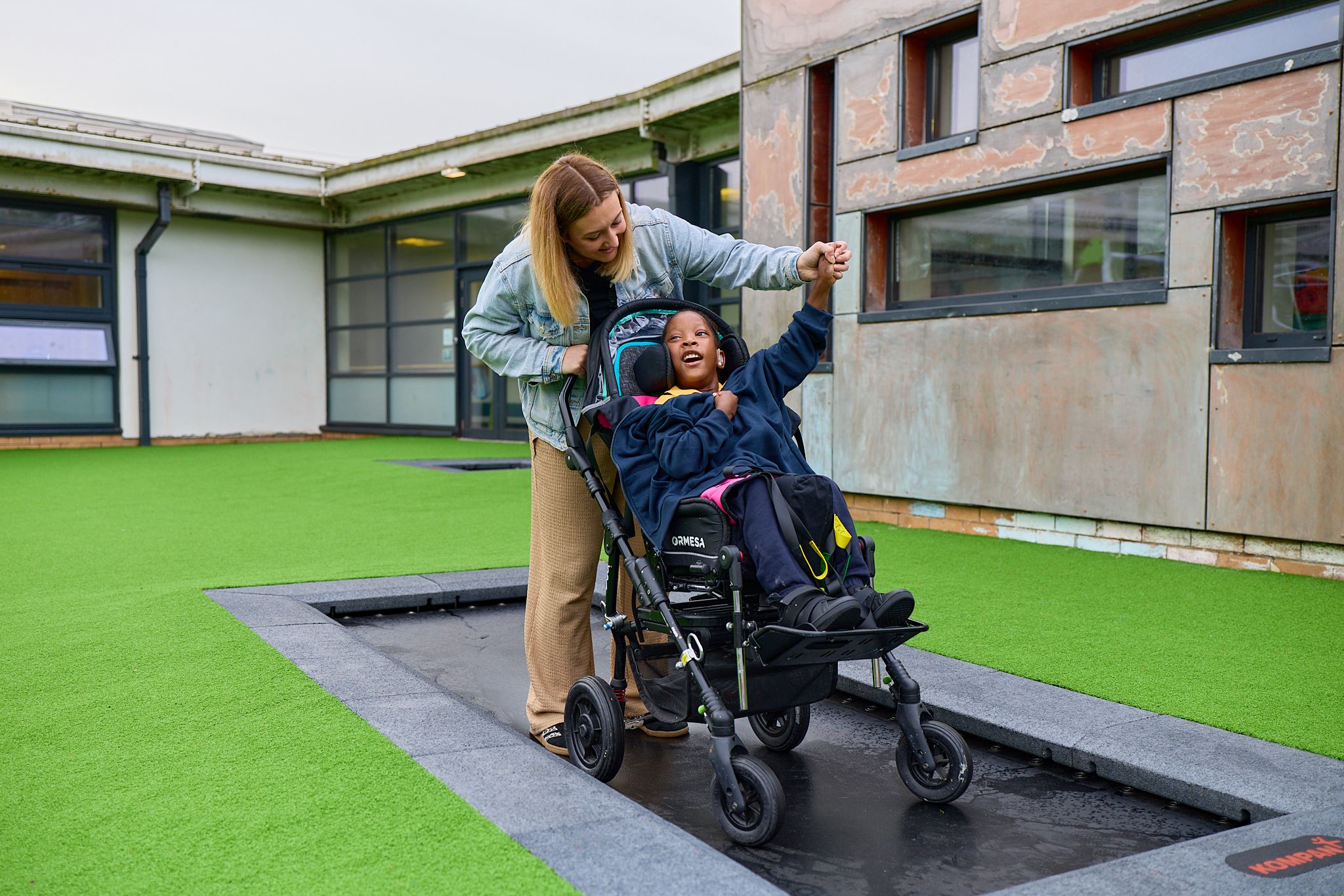 Adult supports child in a wheelchair on an outdoor sunken trampoline beside a modern building with bright green artificial grass