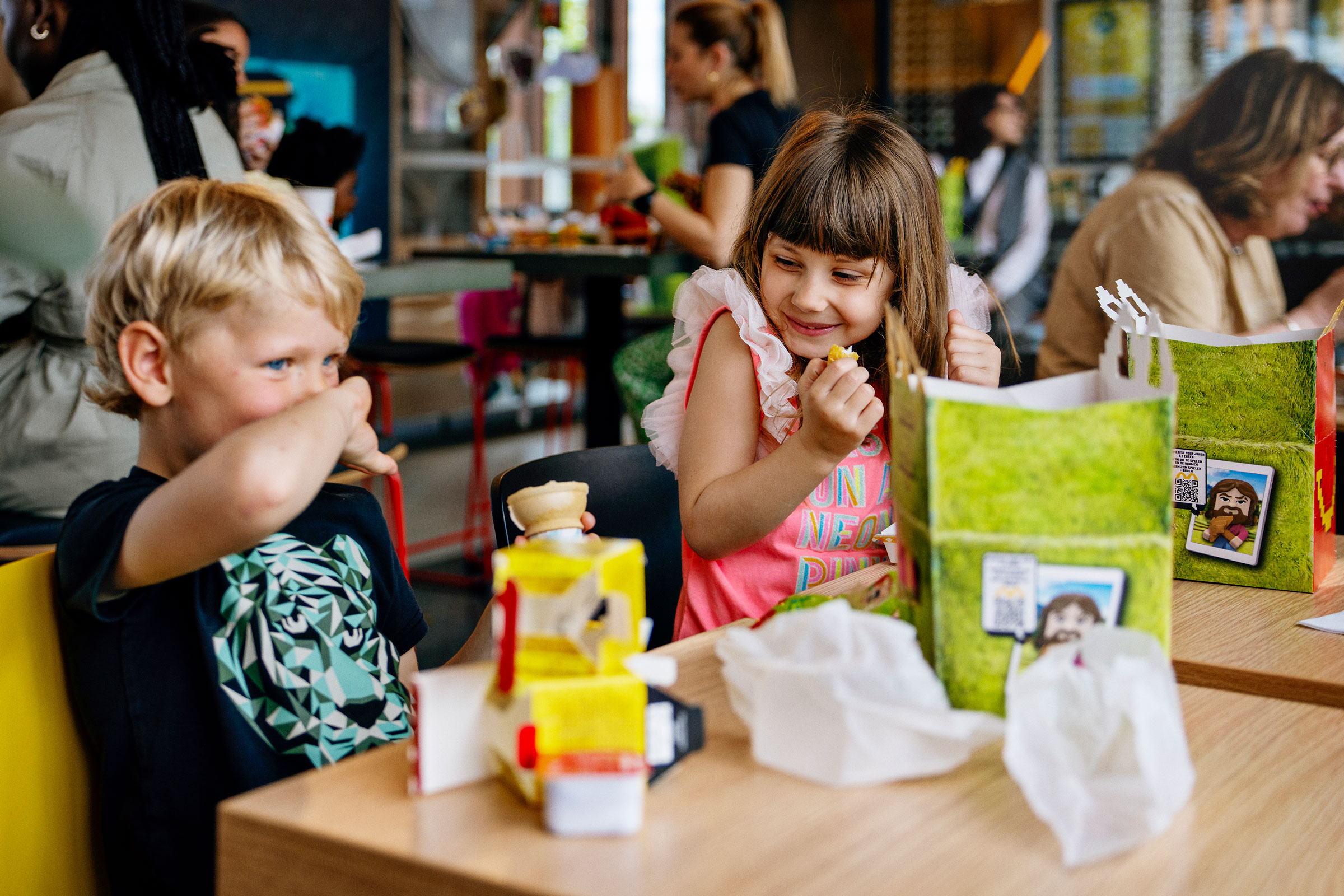 Children sit at a table in a busy cafe, eating from colorful kids’ meal boxes, with adults and other kids in the background.
