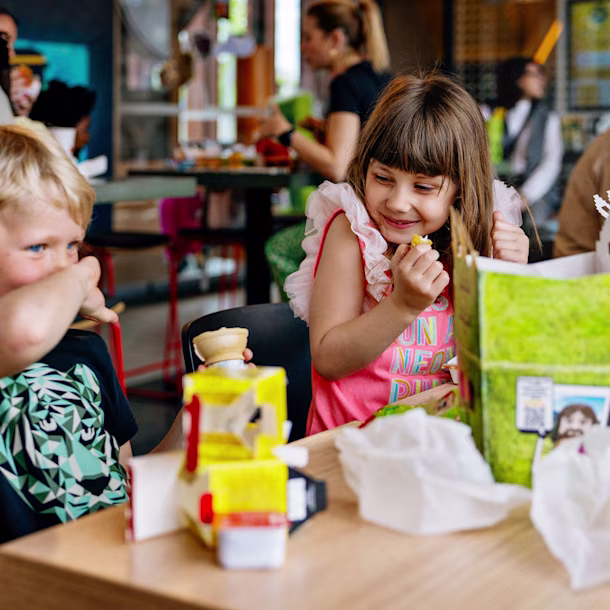 Children sit at a table in a busy cafe, eating from colorful kids’ meal boxes, with adults and other kids in the background.