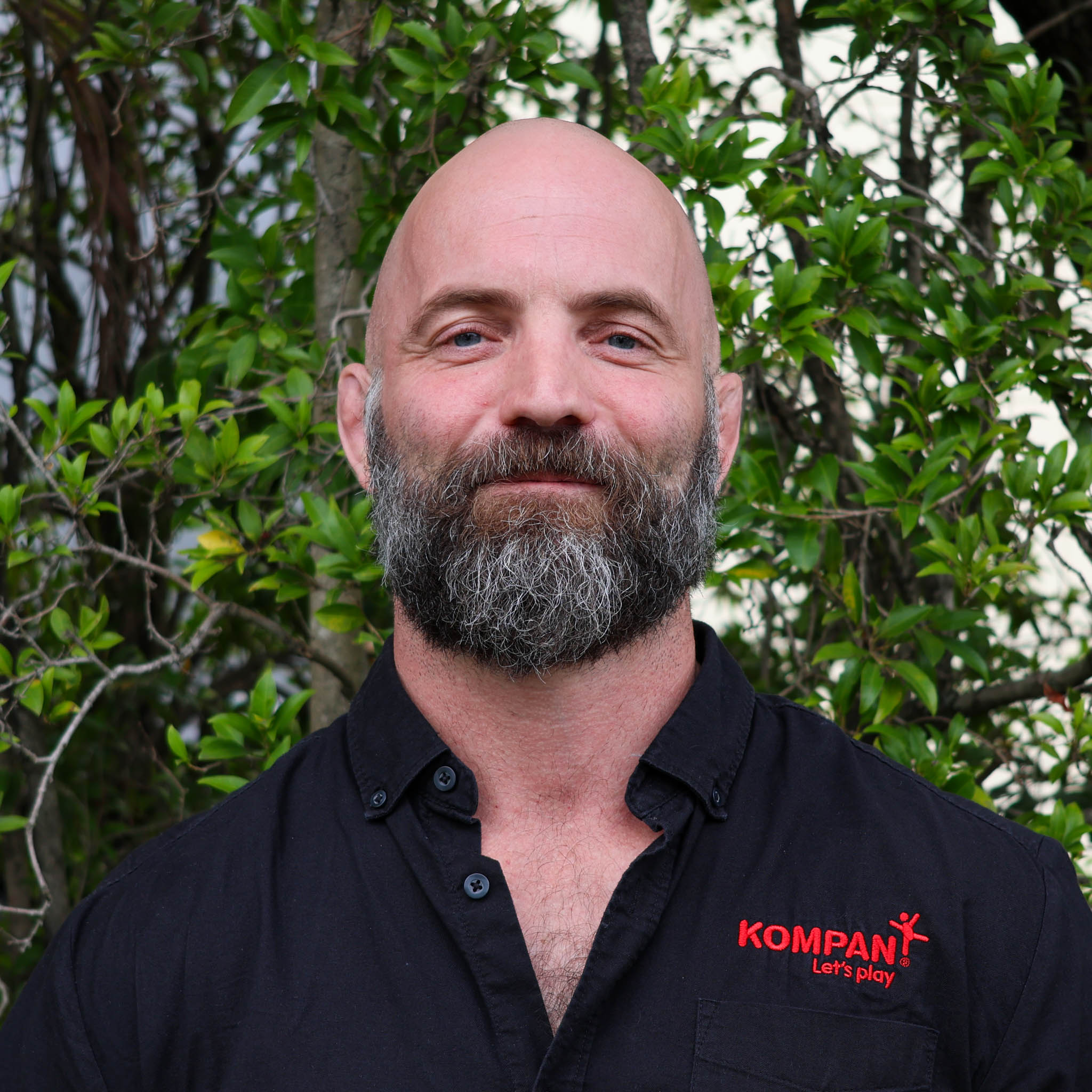 Person with blurred face and beard wearing a black “KOMPAN Let’s play” shirt, standing in front of leafy green trees.