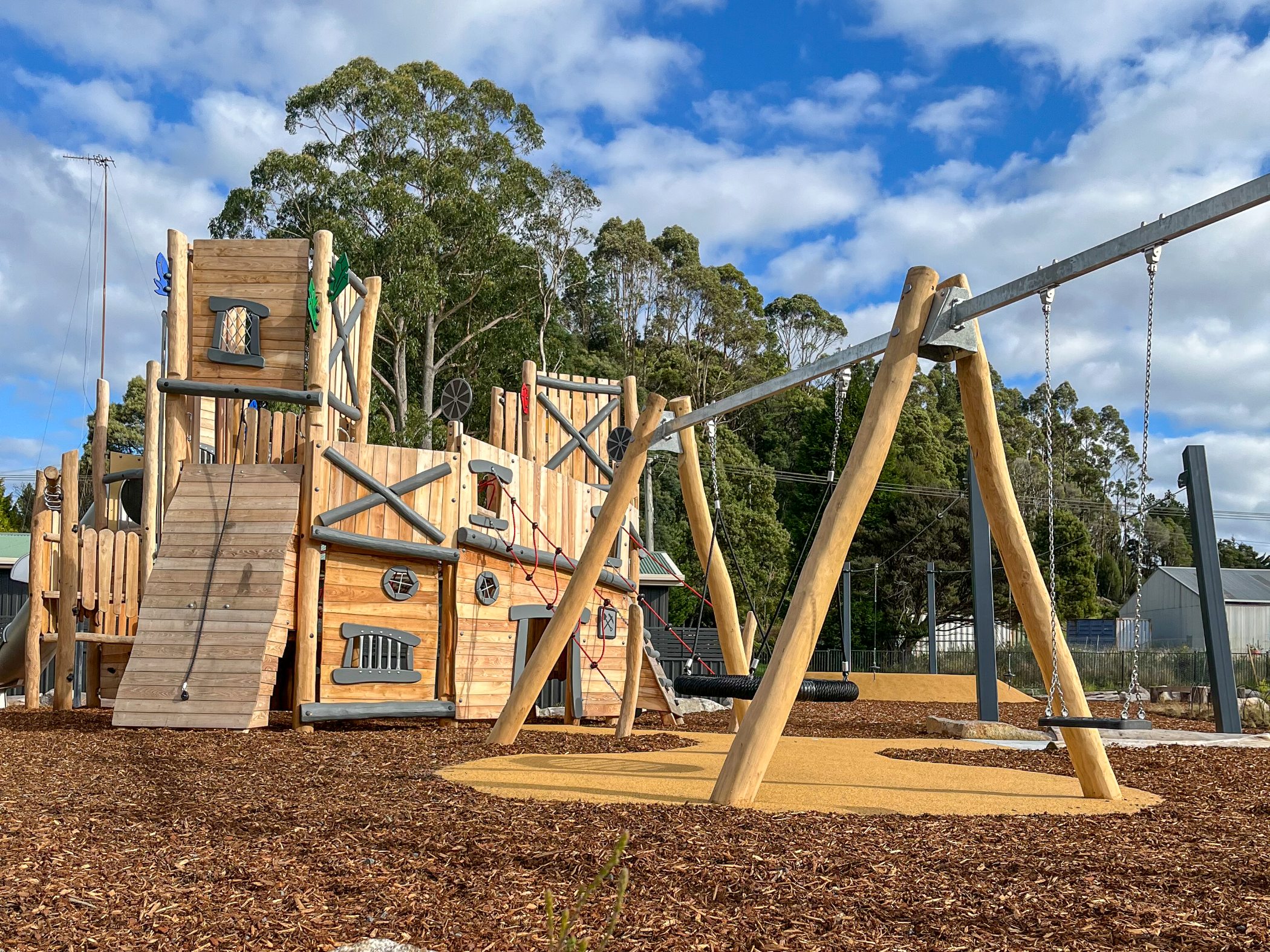 Wooden playground with ship-like climbing structure and swings on mulch ground, set against trees and blue sky with scattered clouds.