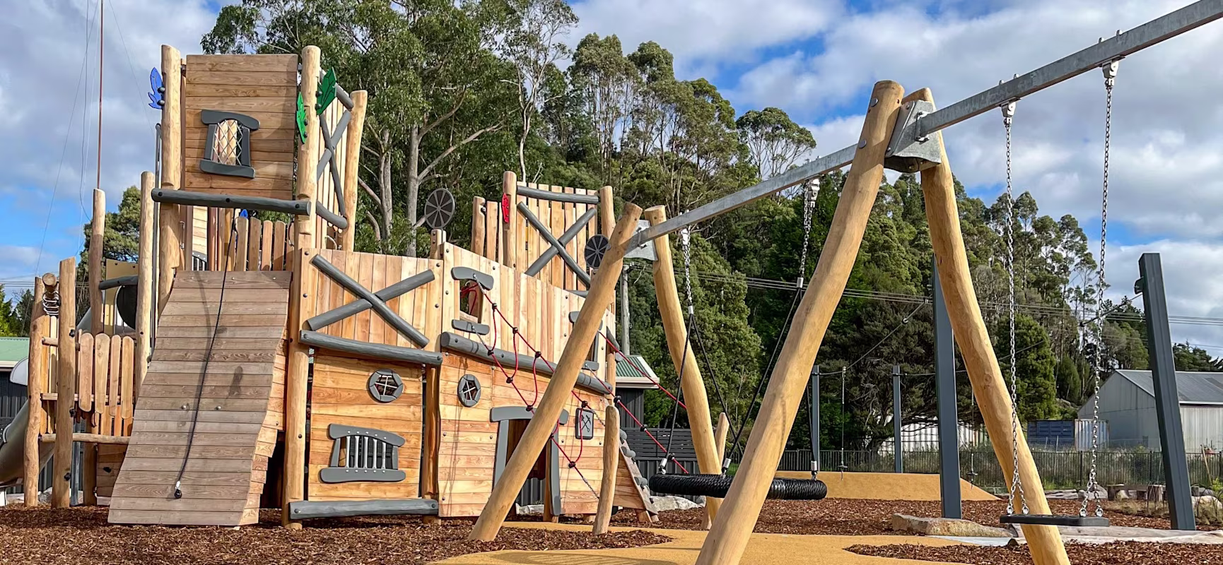Wooden playground with ship-like climbing structure and swings on mulch ground, set against trees and blue sky with scattered clouds.