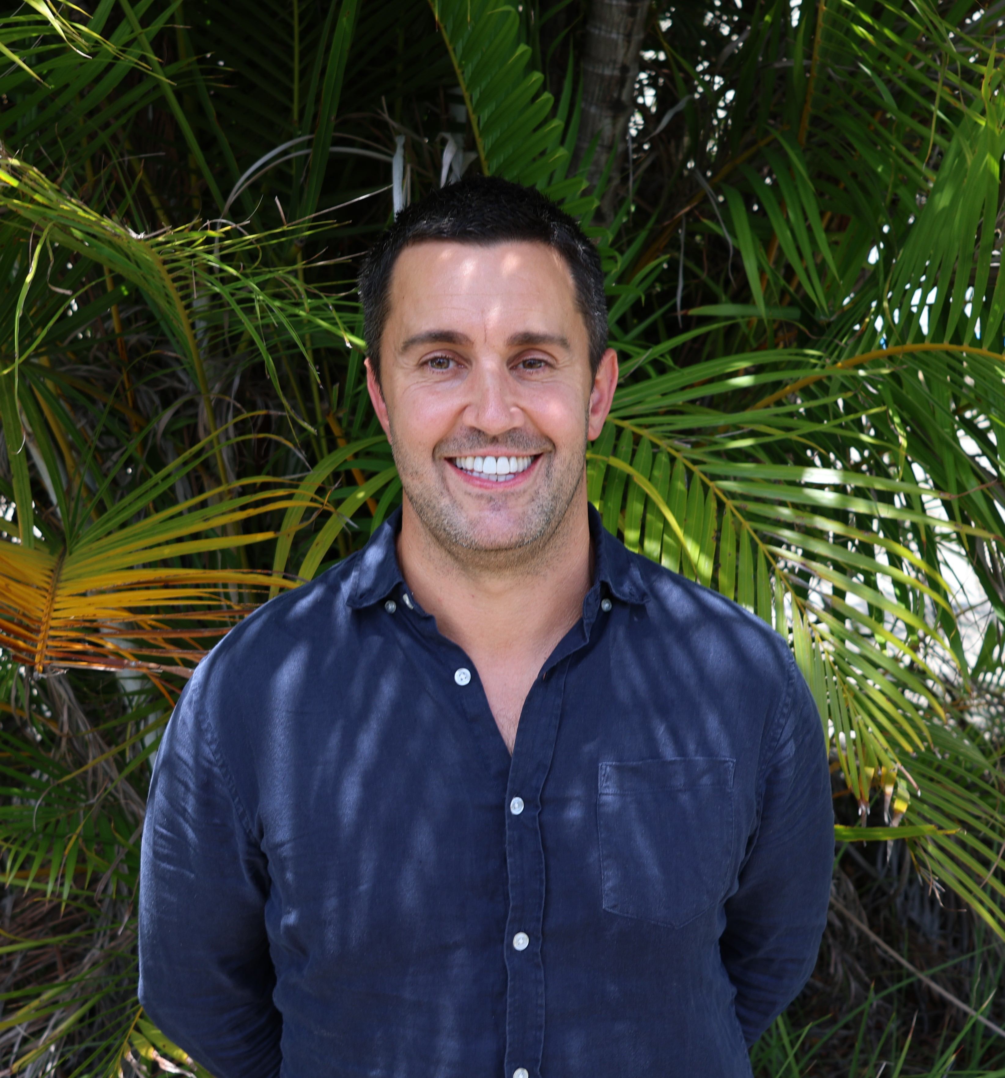 Person with blurred face in a navy shirt standing in front of dense green palm leaves in bright outdoor light