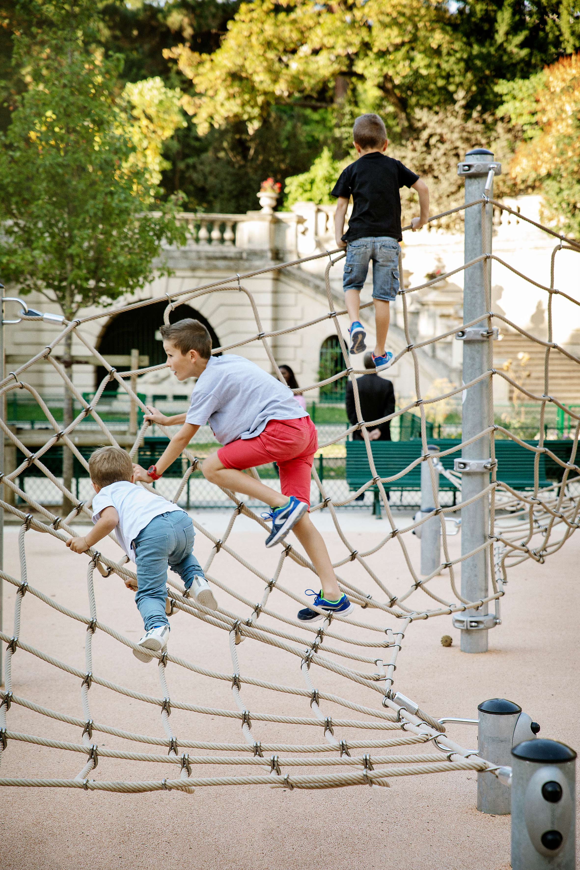 Three children climb on a rope net playground structure in a sunny park with trees and a stone building in the background.