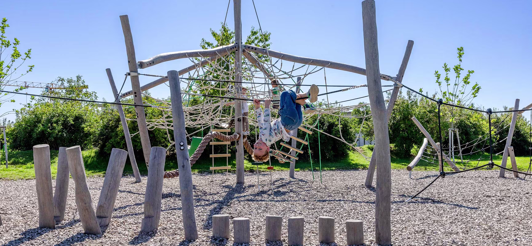 Child hanging upside down on a wooden rope climbing frame in a sunny playground, surrounded by mulch ground and green trees.