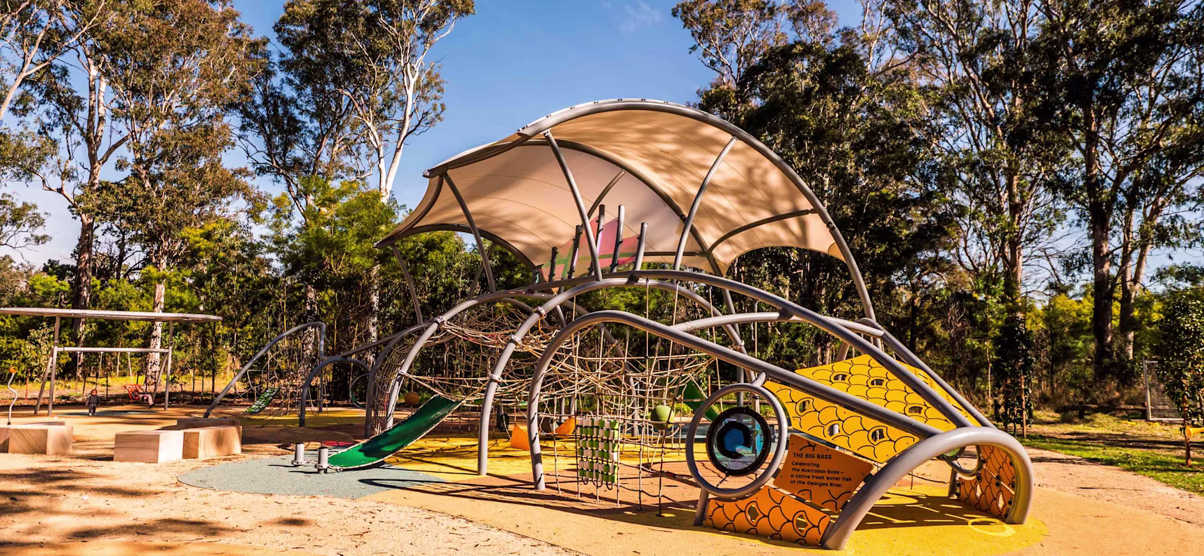 Modern playground with curved climbing frame, slide and shade canopy, set on soft ground among tall trees under clear blue sky