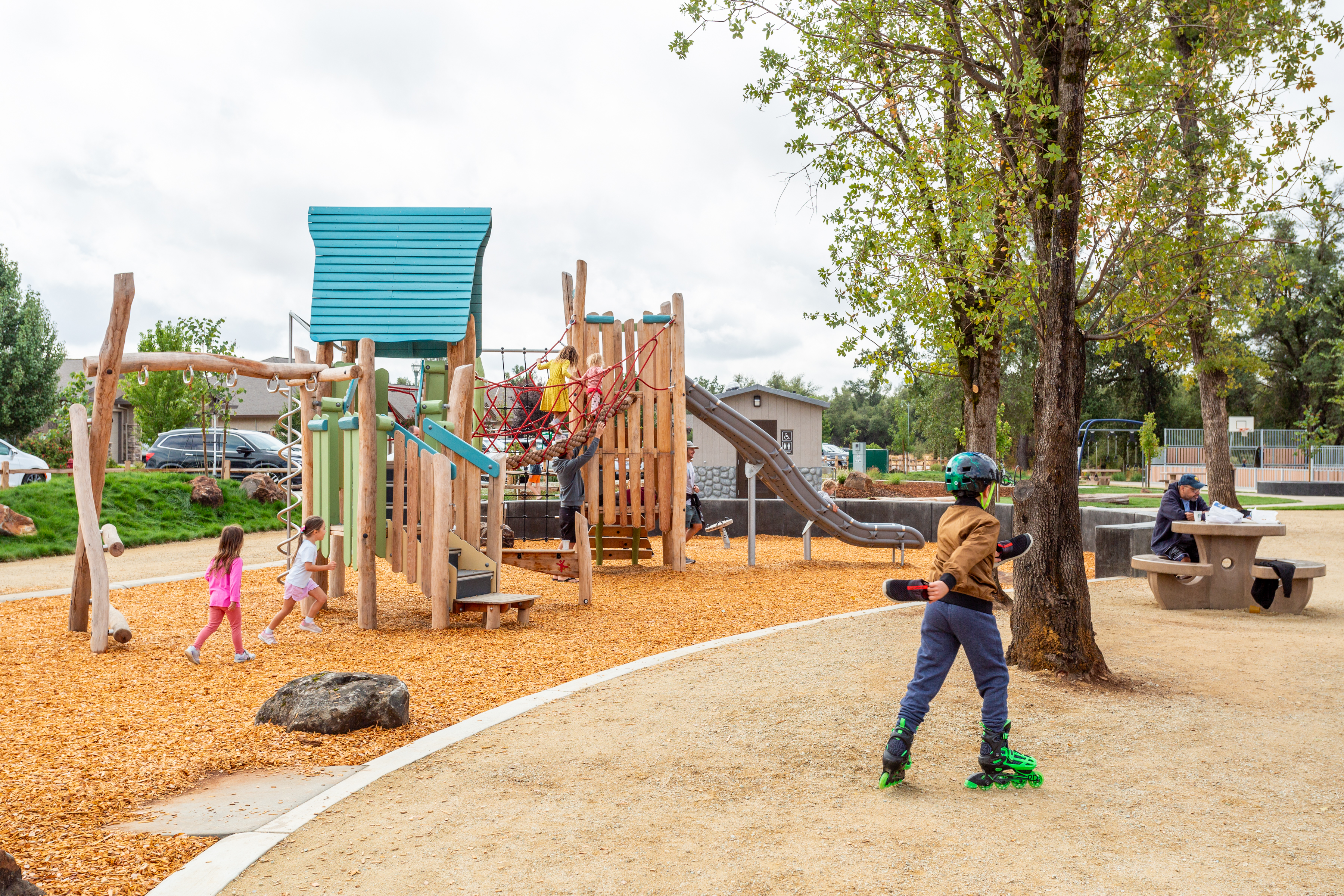 Kids playing on natural commercial playground equipment at community park in Sacramento, California as a child skates by in the foreground