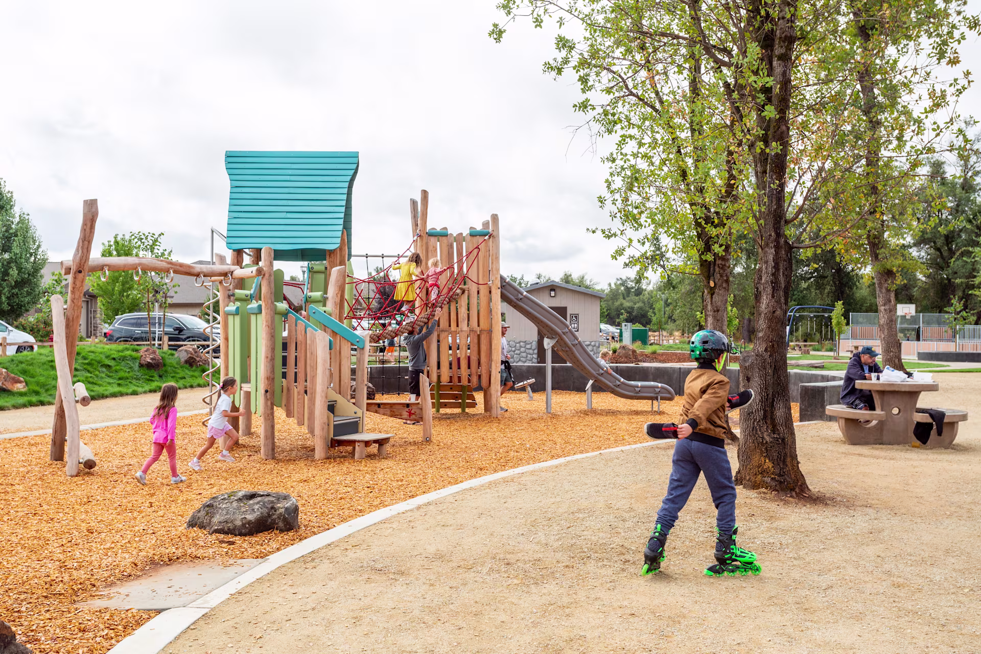 Kids playing on natural commercial playground equipment at community park in Sacramento, California as a child skates by in the foreground