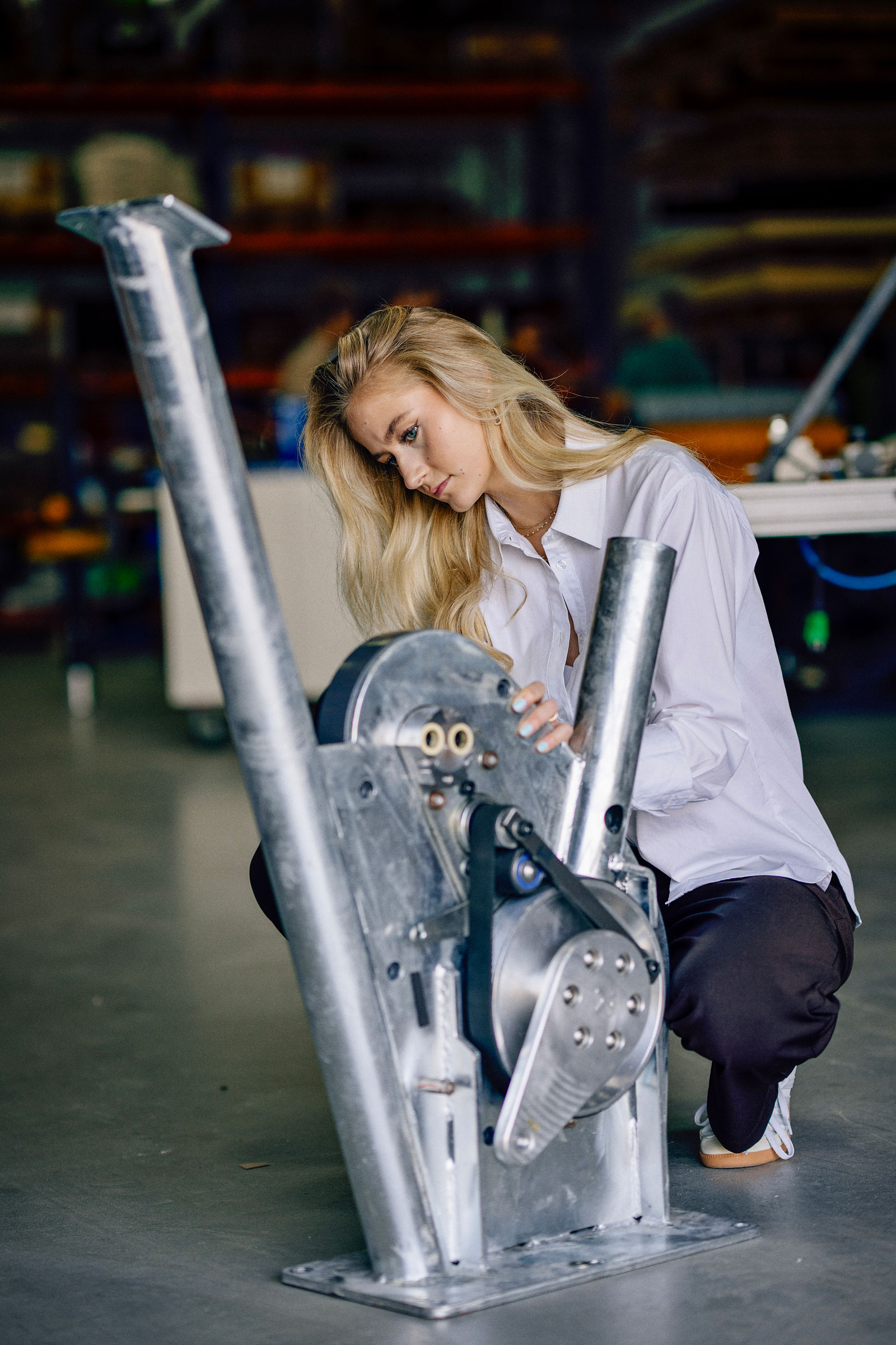 Person in a white shirt crouches in a workshop, examining a large metal mechanical device with belts and pulleys.