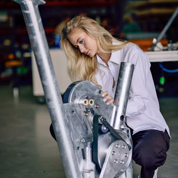 Person in a white shirt crouches in a workshop, examining a large metal mechanical device with belts and pulleys.
