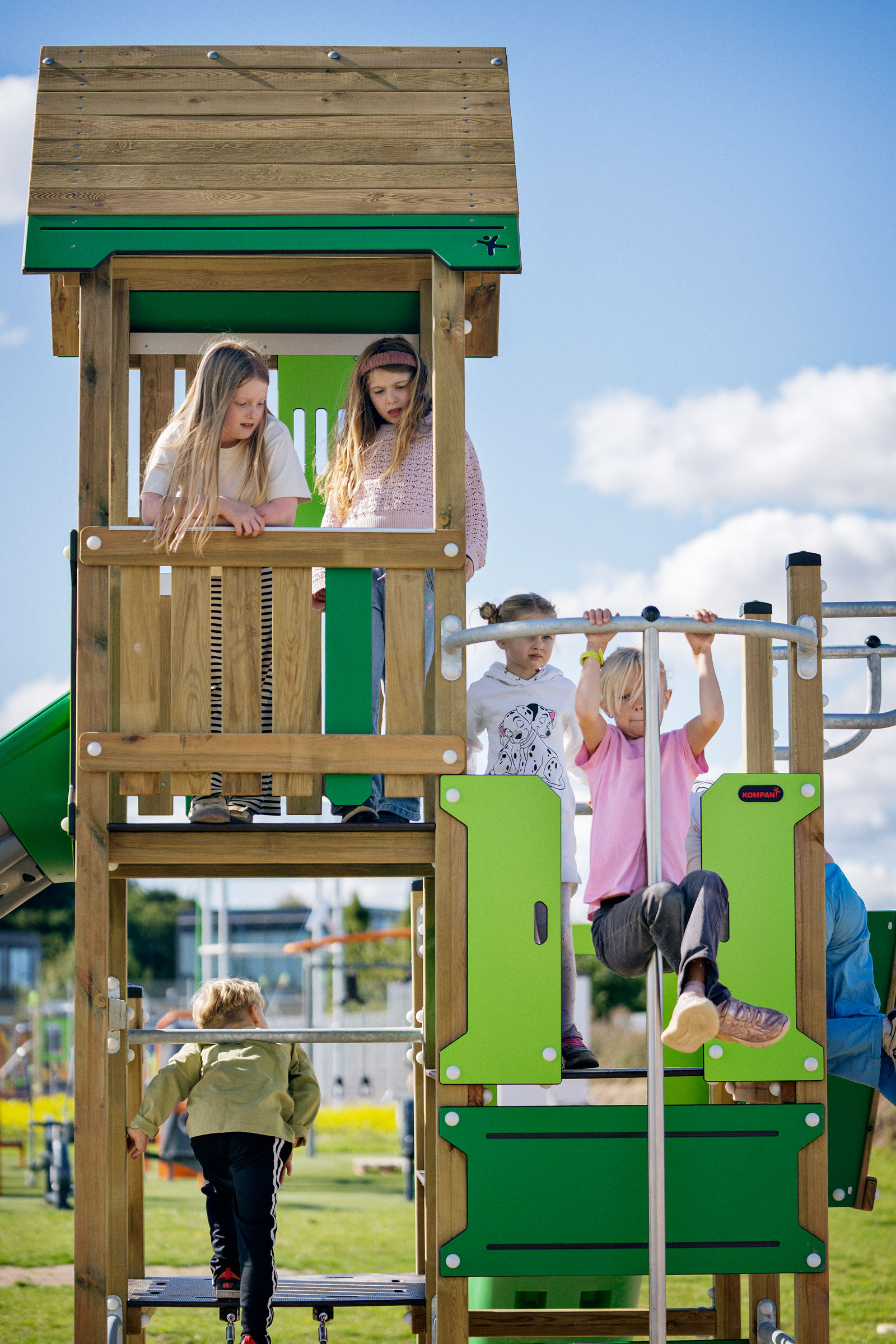 Children climbing and hanging on a wooden and green playground structure on a sunny day with blue sky and clouds