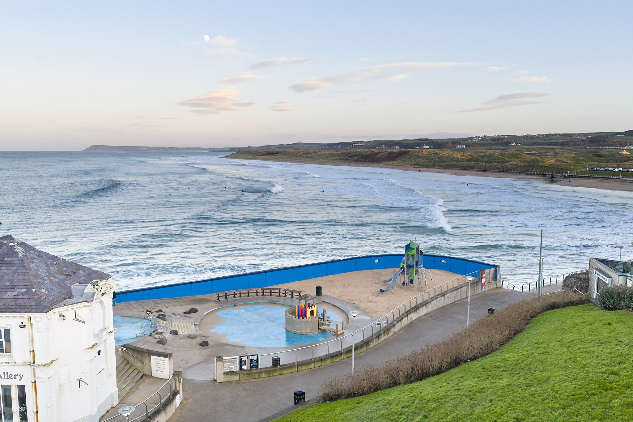 Seaside view with waves, a sandy playground and paddling pool behind blue fencing, beside a promenade and grassy slope.